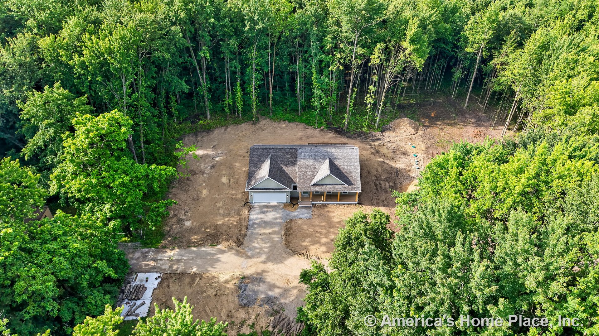 Single-story exterior featuring a covered front porch, attached two-car garage, gable roof, and unfinished driveway, set on a wooded lot surrounded by dense forest.