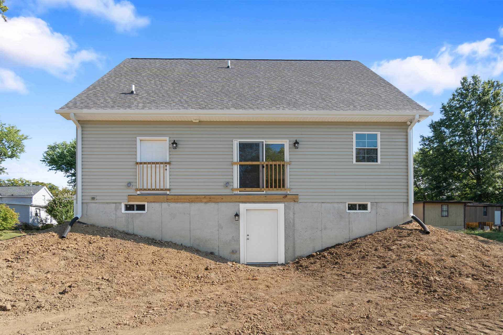 Two-story home with white siding, wooden railings, and large windows, set against a grassy hill and mature trees under a cloudy sky