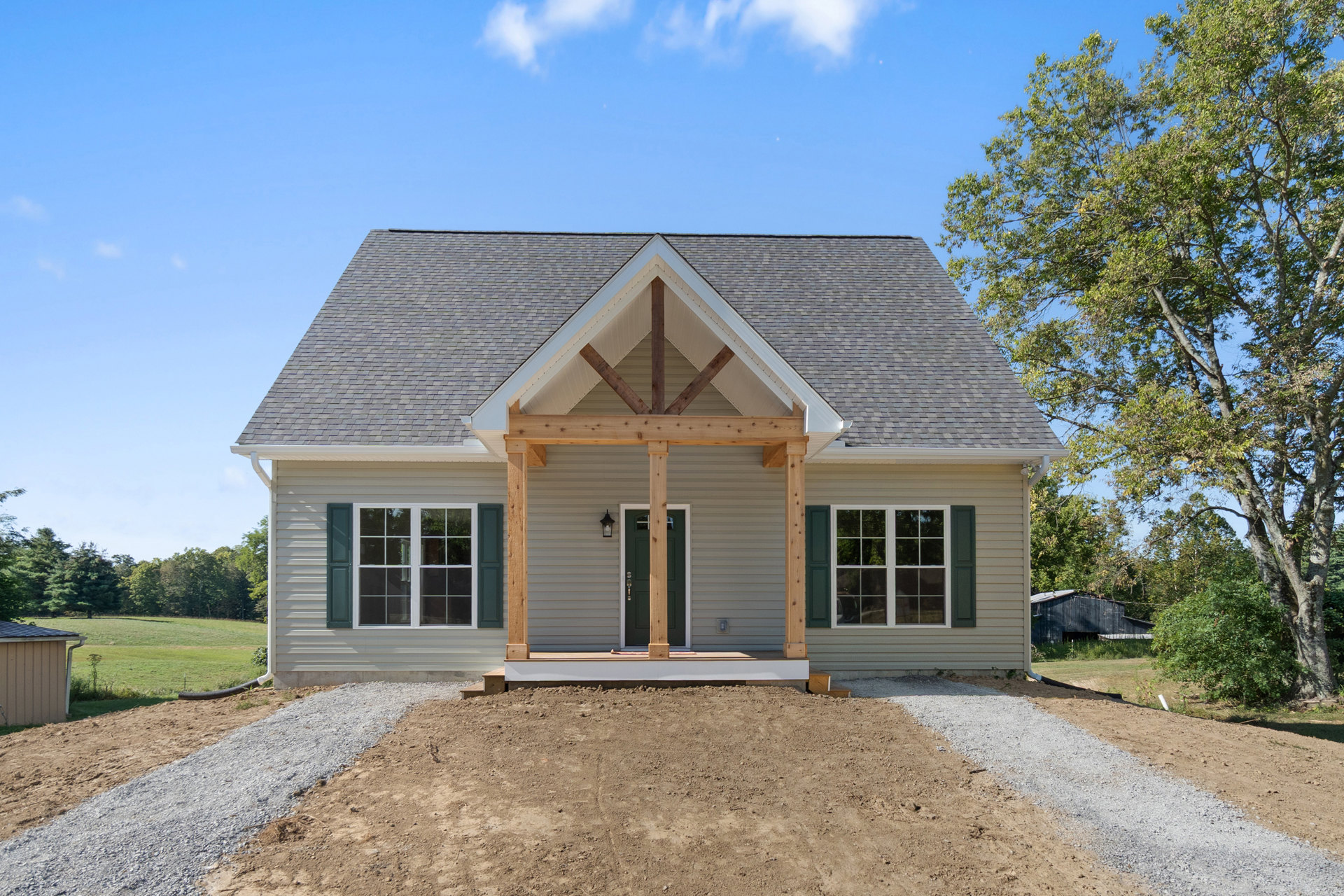 Wooden porch with vertical posts, green front door, multi-pane window, dirt ground, and leafy trees in the background