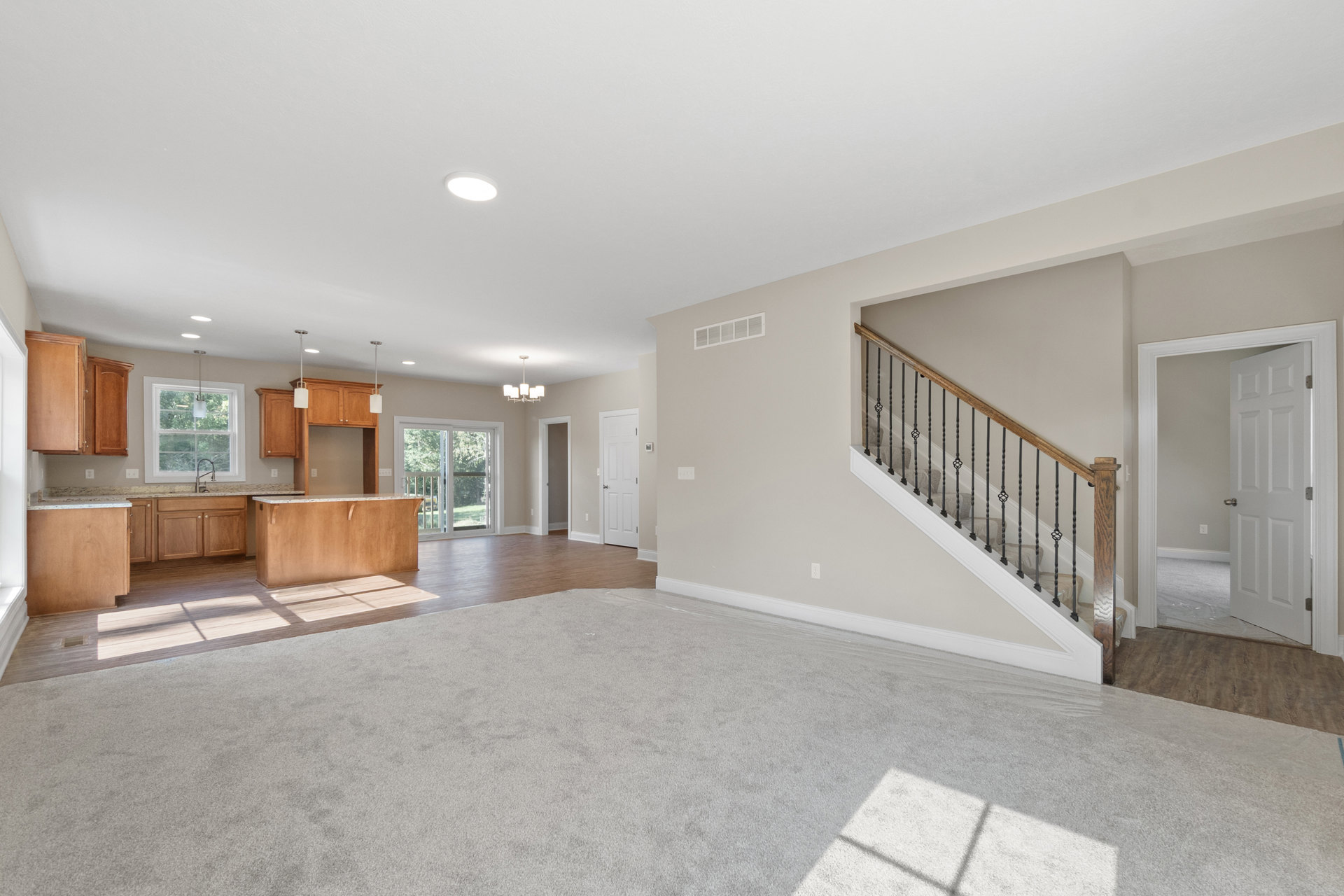 Open living room with laminate flooring, white walls, staircase with wooden treads and metal railing, adjacent kitchen featuring light fixtures above sink, sliding glass door, and