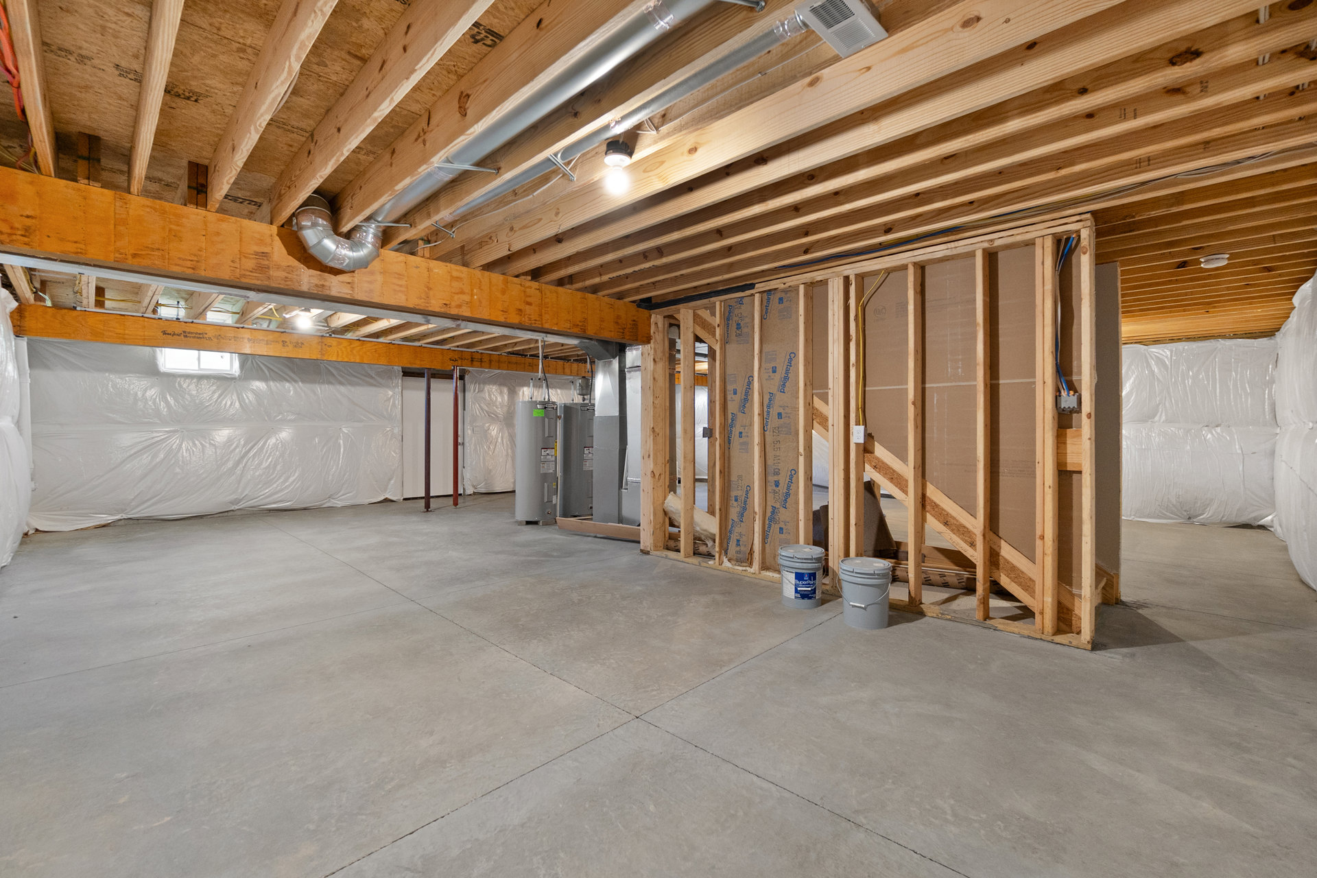 Open room with exposed wood ceiling beams, unfinished concrete floor, paint bucket on floor, and partially constructed wood plank walls