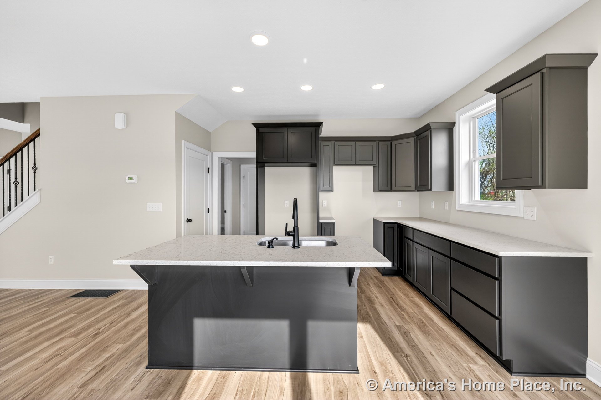 Dark shaker cabinets and white quartz countertops line the kitchen, centered by a large island with an undermount sink; recessed ceiling lights illuminate wood-look vinyl flooring
