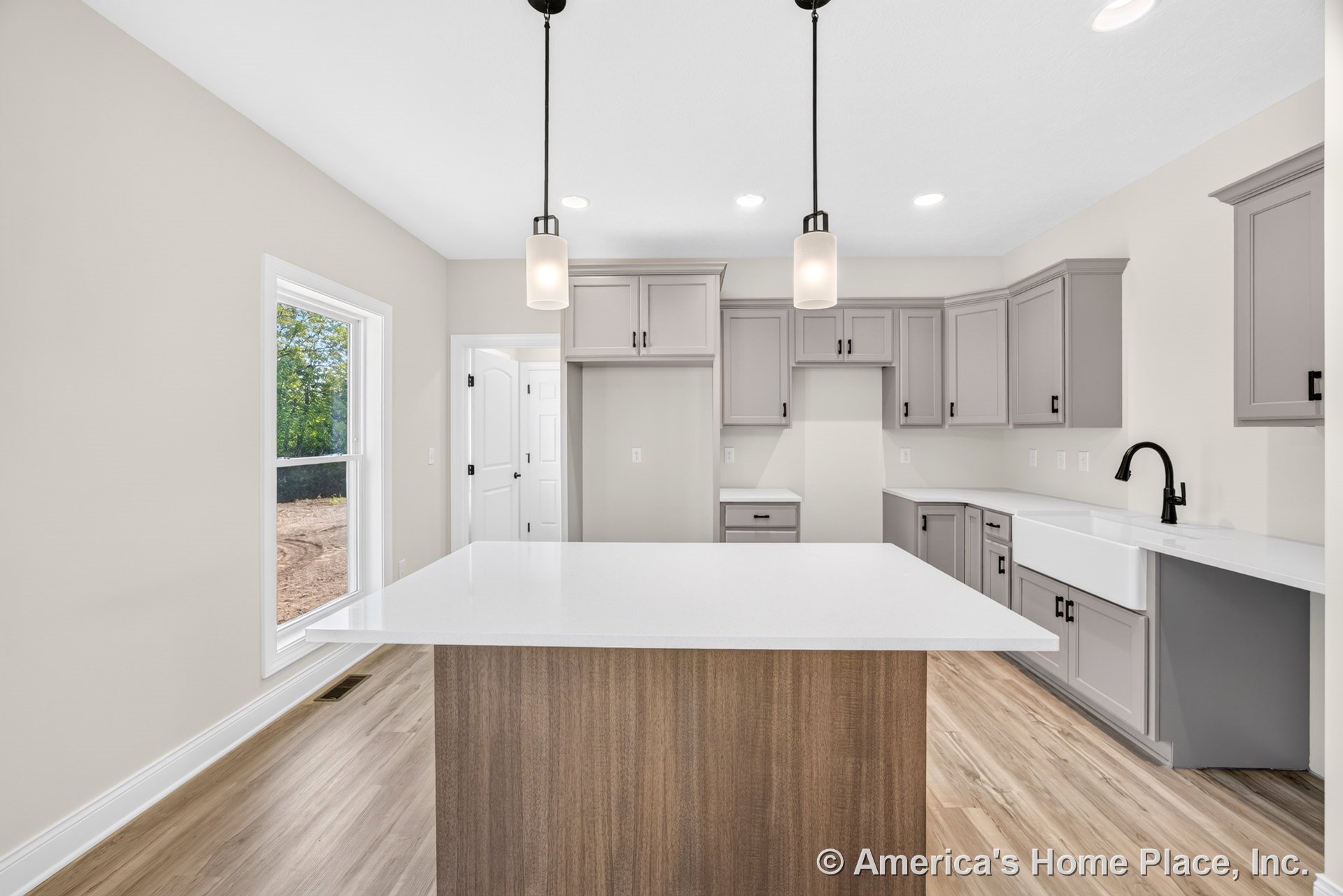 Light gray shaker cabinets, large central island with white quartz countertop, farmhouse sink with black faucet, pendant and recessed lighting, wood-look flooring, expansive window