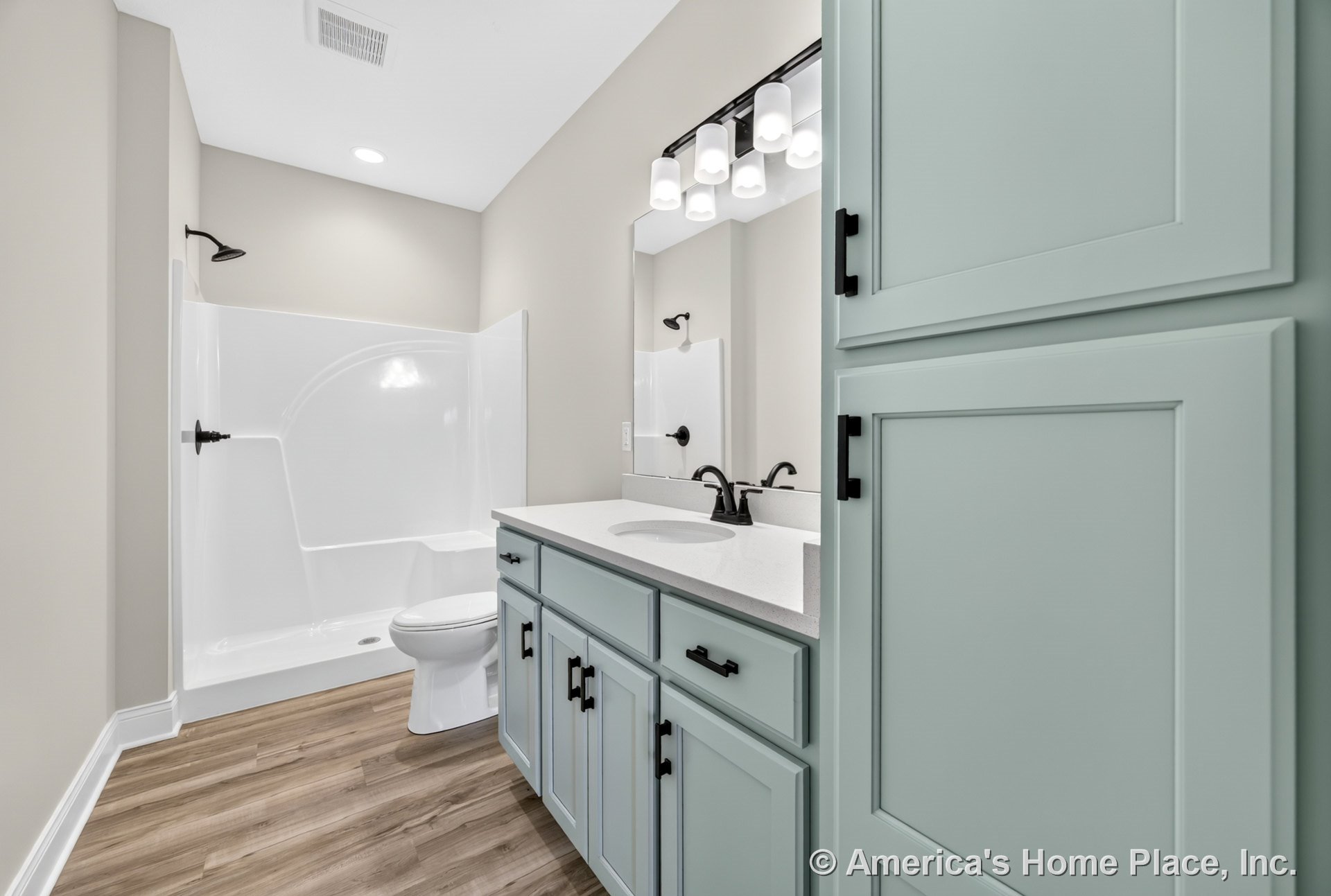 Light blue built-in cabinetry with white solid surface countertop, walk-in shower featuring black fixtures, wood-look flooring, modern vanity lighting, large mirror, and built-in