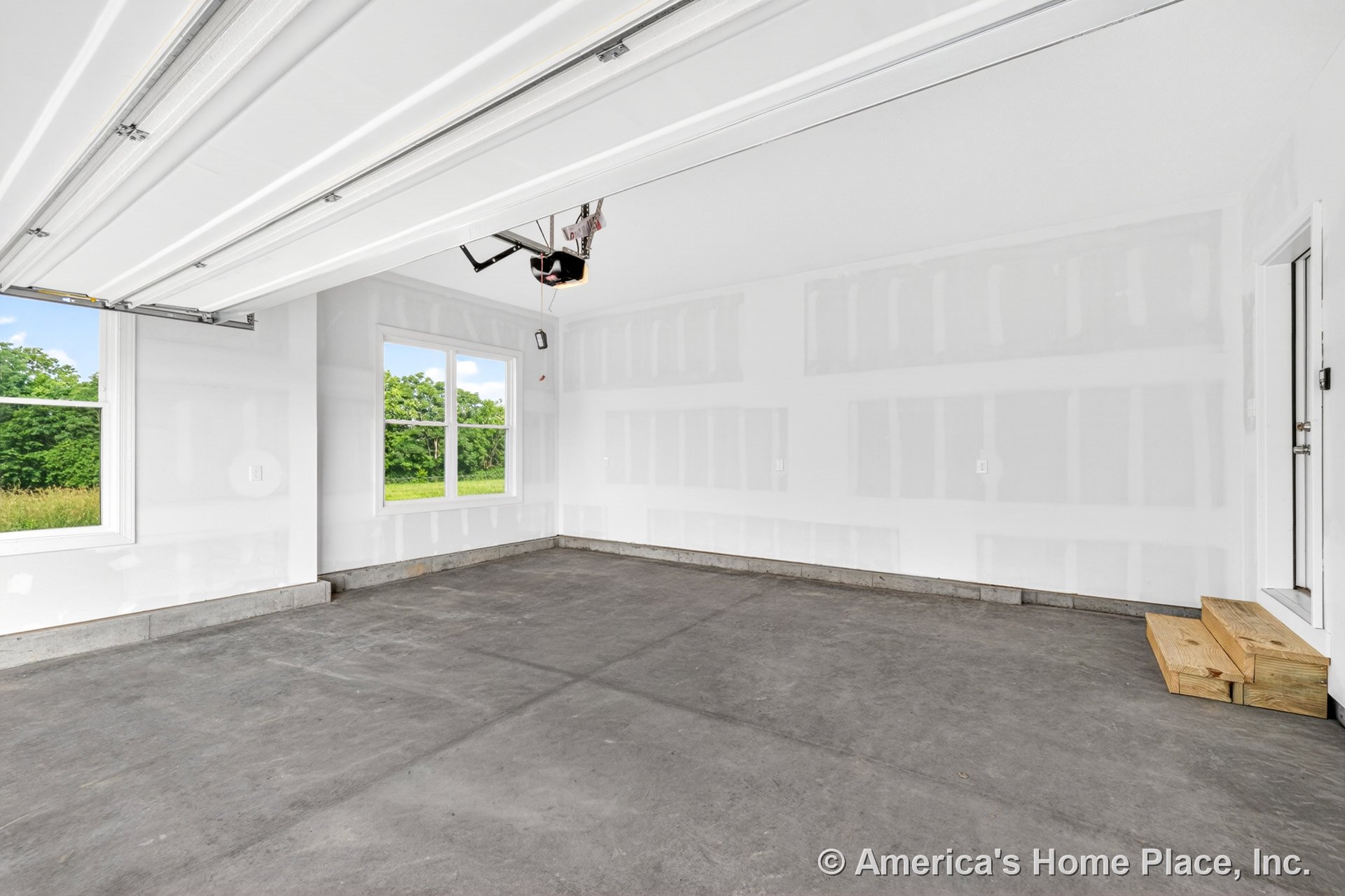 Unfinished drywall garage with concrete slab floor, double window providing natural light, garage door opener, white ceiling, wooden entry steps, wall outlets, and exterior door