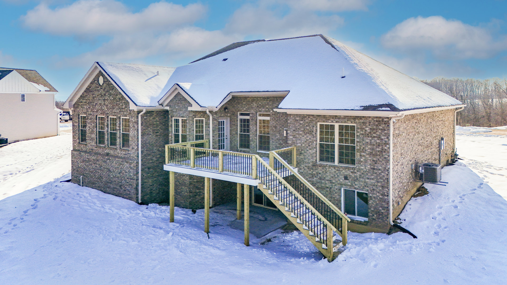 Two-story house with white-framed windows, wood deck featuring railing and stairs, snow covering patio and roof, winter landscape with cloudy sky and mountain in background