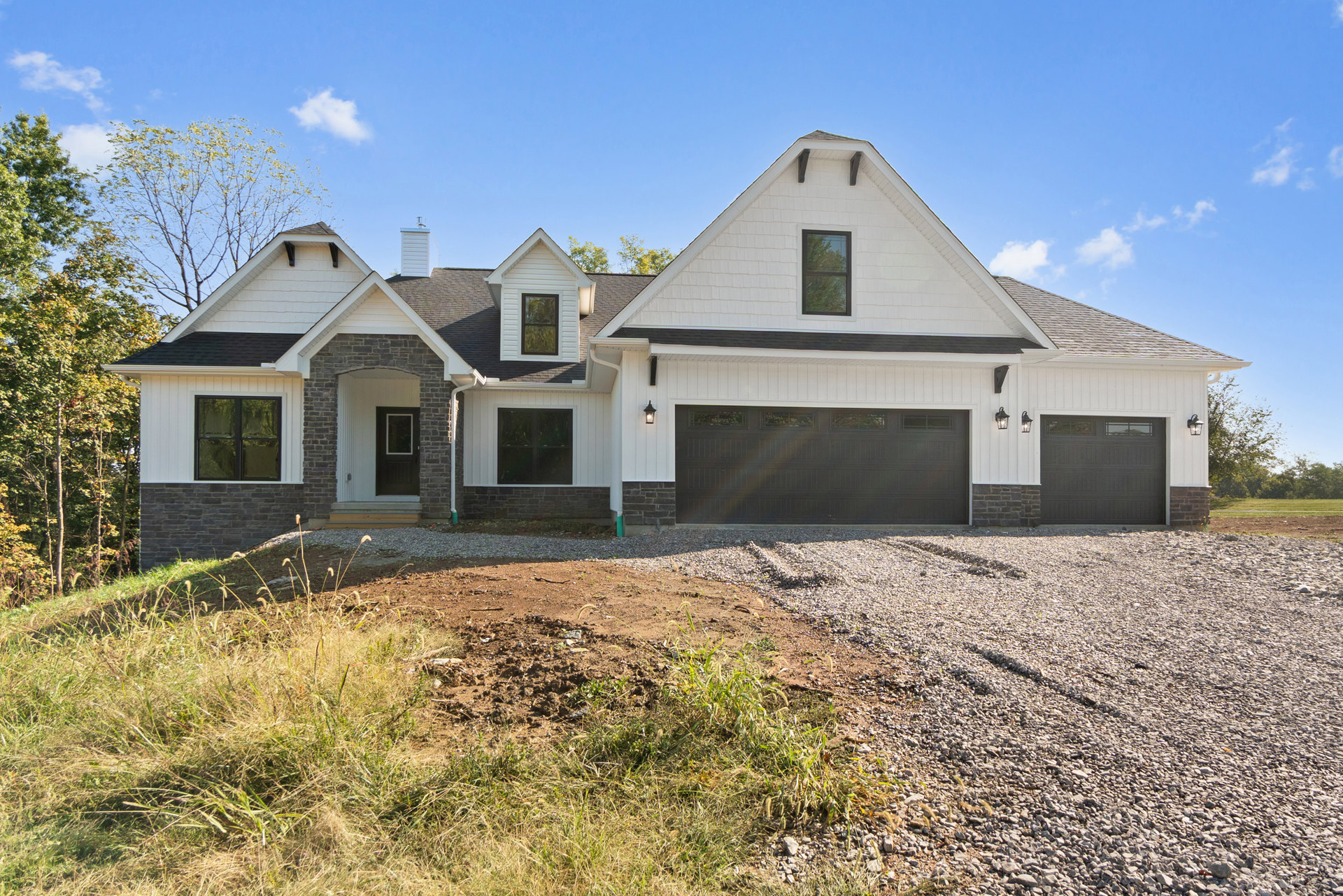 Modern home exterior with black-framed windows, black garage door with upper windows, concrete driveway, dirt landscaping patch, and trees reflected in glass