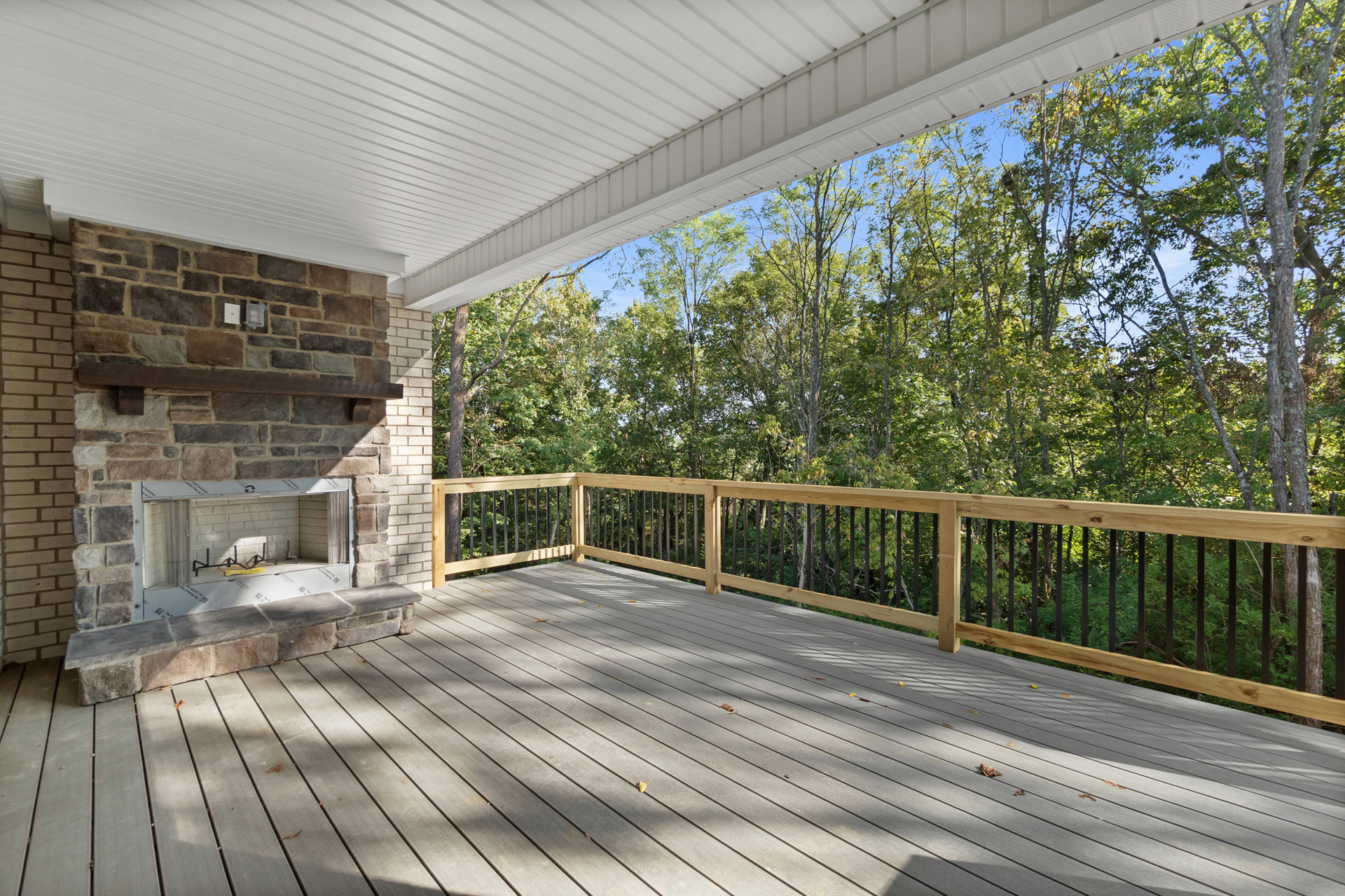 Wooden deck with white brick fireplace, black metal firewood holder, railing, and leafy trees in the background