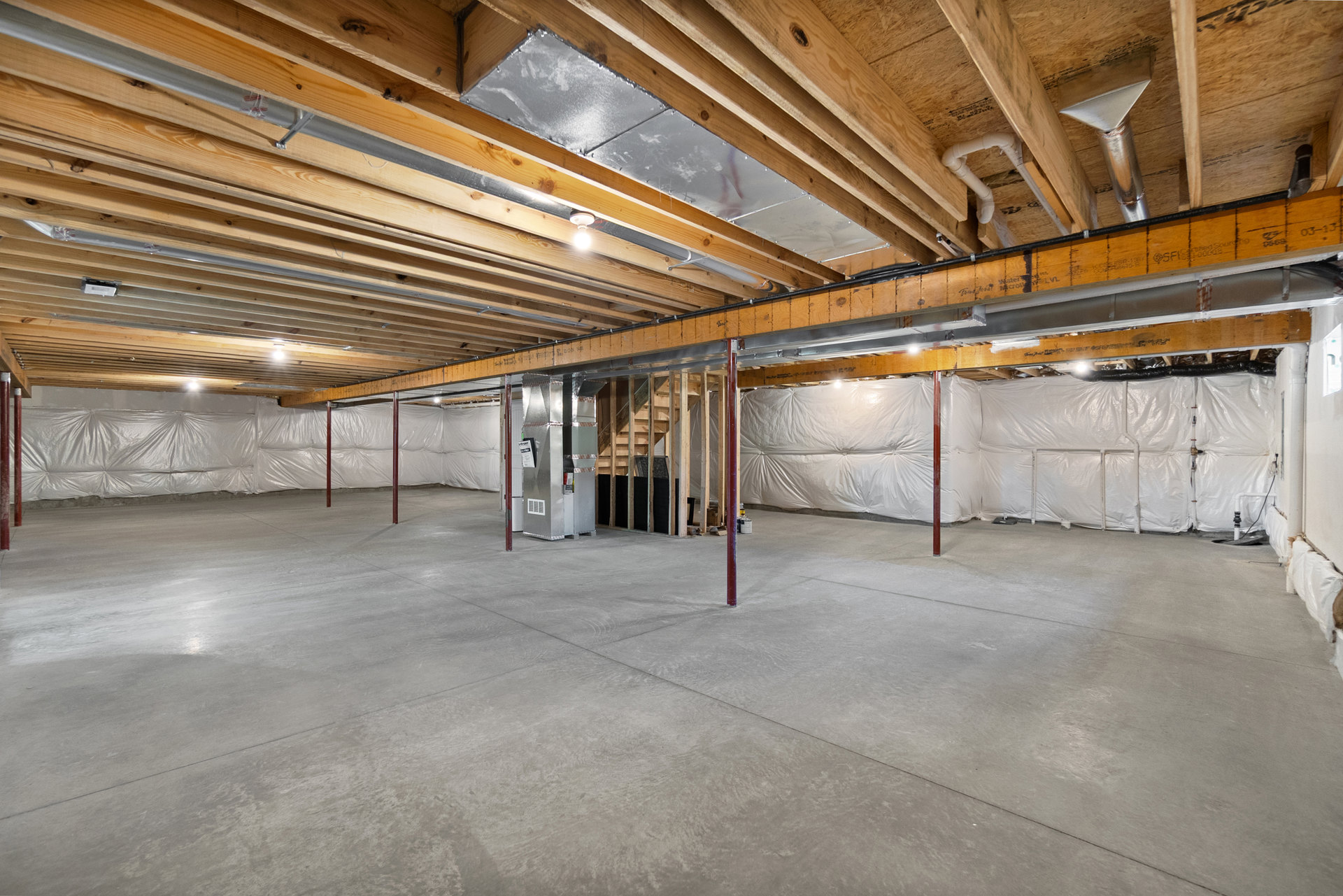 Concrete floor with exposed wooden ceiling beams, red support pole, metal pipe with triangular top, single light bulb hanging from ceiling, unfinished walls.