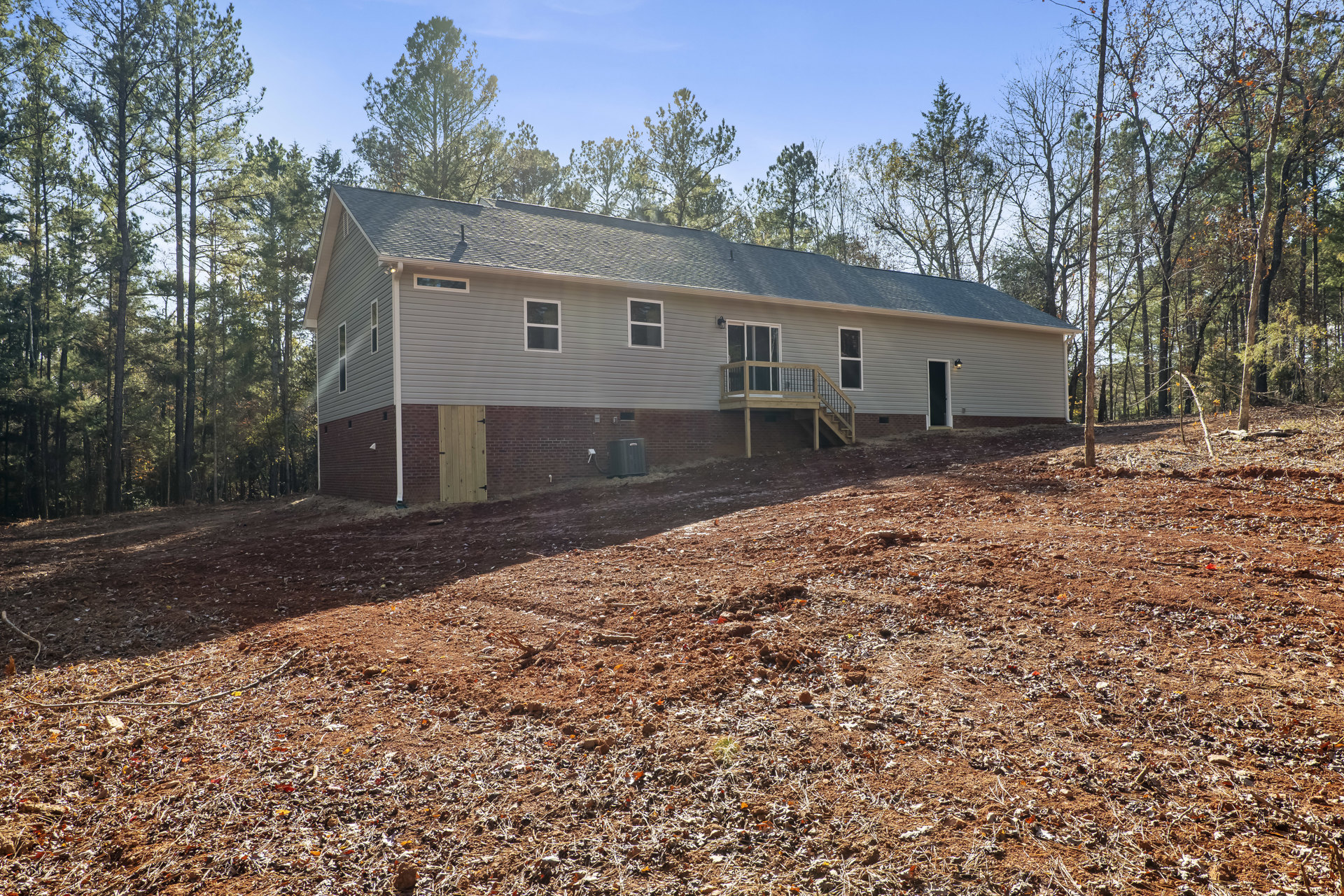 Brick house with wooden deck and glass door, spacious grassy yard with trees, autumn foliage, outdoor stairs leading up a dirt hill, black utility box near brick wall, wooden door