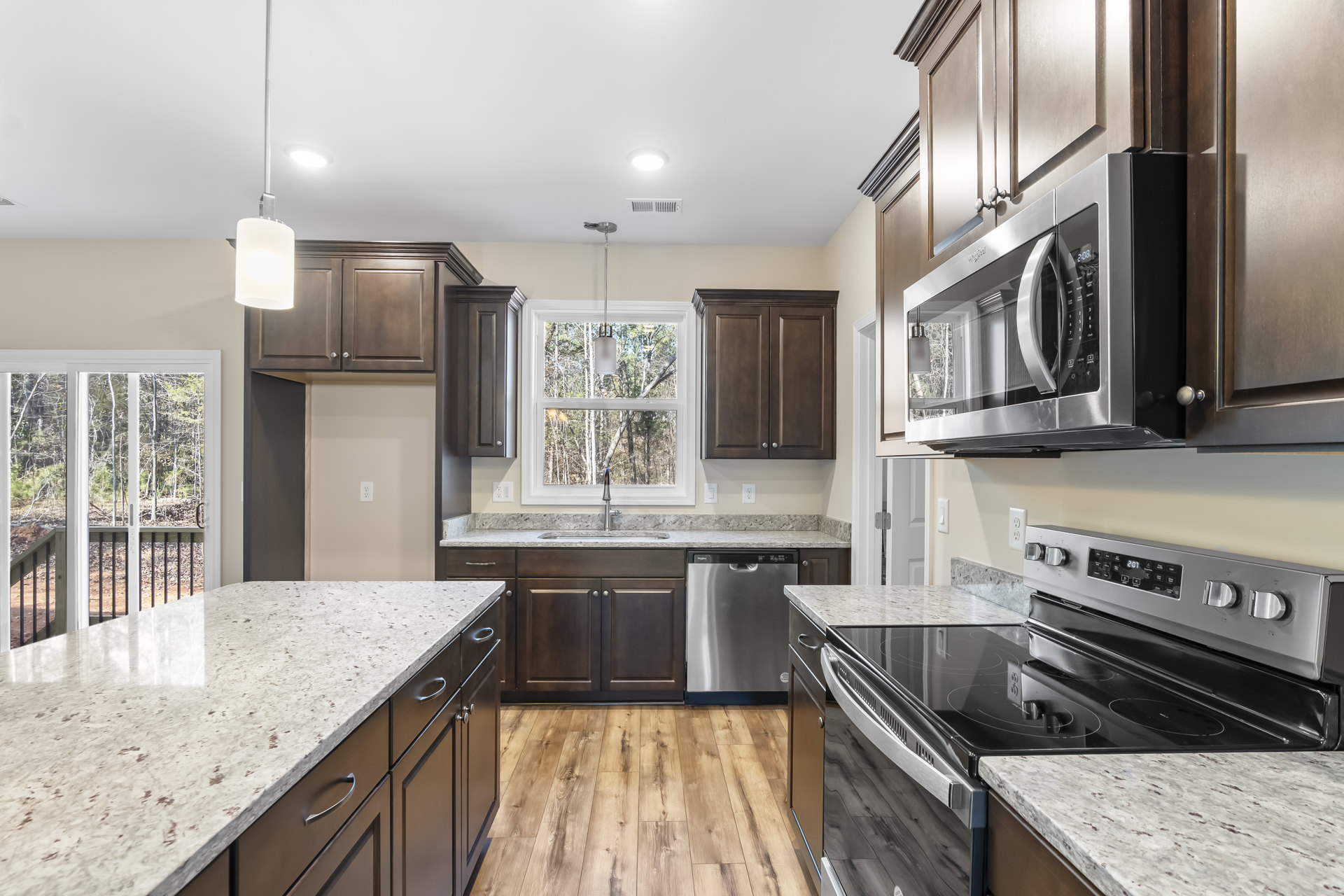 Granite countertops, stainless steel appliances, brown cabinetry, microwave with glass door, window and sliding glass door overlooking trees