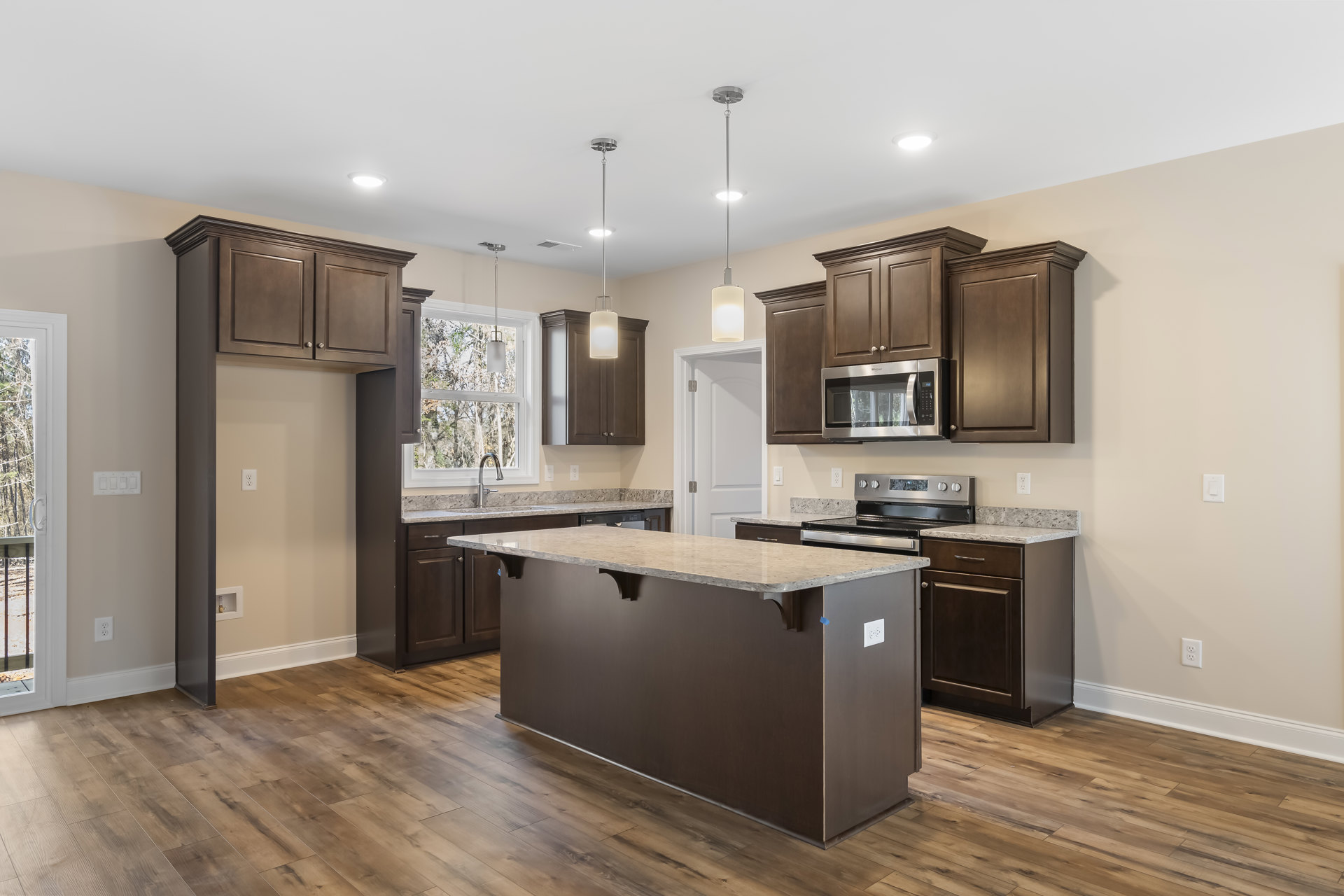Spacious kitchen featuring a large granite island, wood flooring, brown cabinetry, built-in microwave, white walls, and pendant lighting.