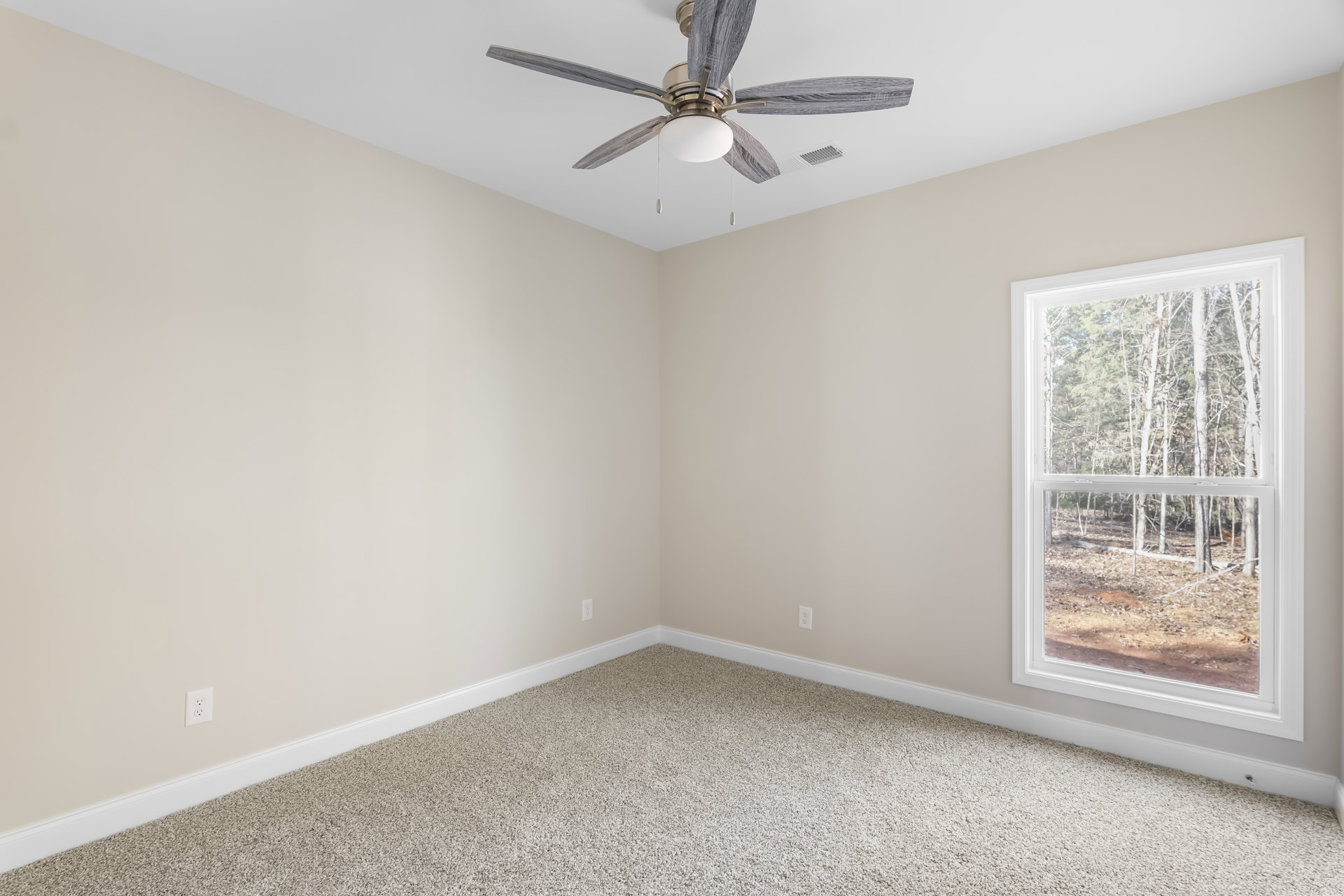 Neutral-toned carpeted room with white walls, ceiling fan with light fixture, large window overlooking trees, electrical outlet visible near baseboard