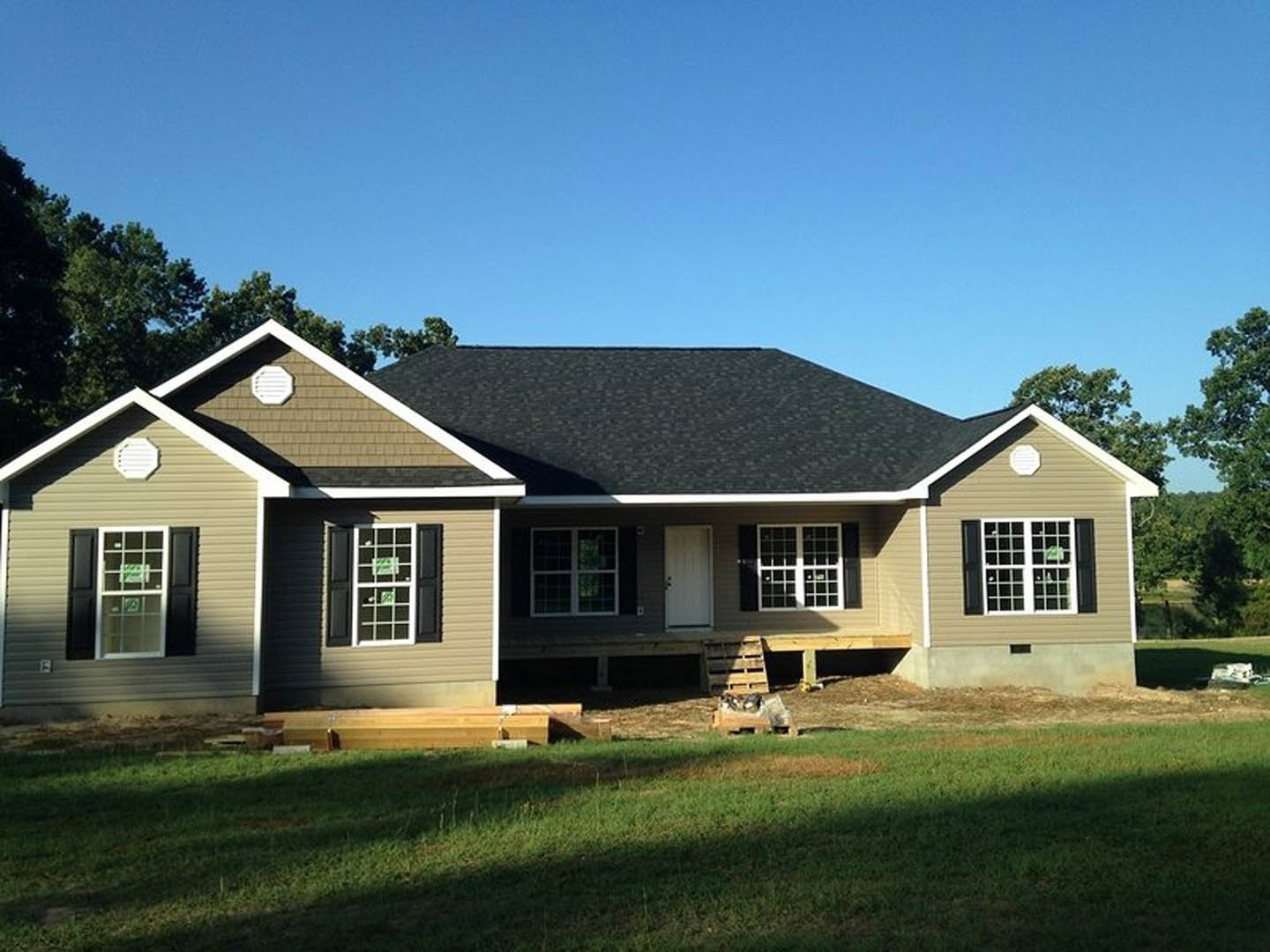 Partially built house with exposed wooden framing, covered porch, white-framed windows, brick wall with a white sign, surrounded by green lawn and trees under a blue sky
