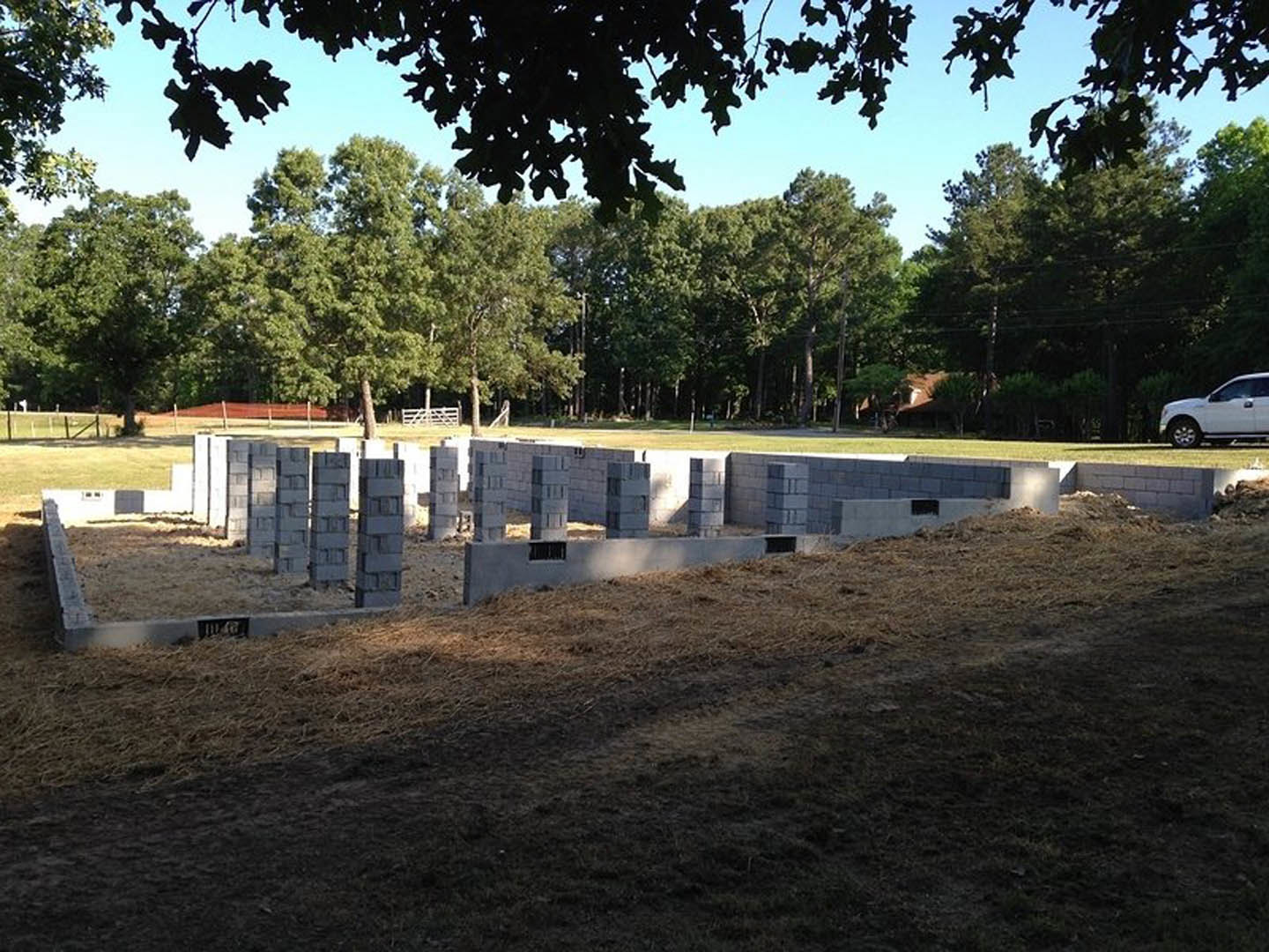 Modern custom home with light-colored exterior, partially built foundation, wooden fence, mature trees, and grassy yard.