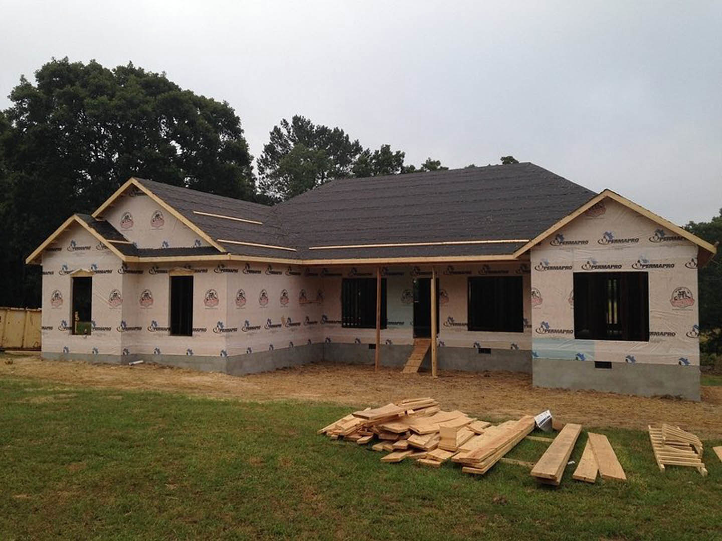 Framed house under construction with exposed wood planks, unfinished wooden staircase, pile of lumber on grassy lot, window installation, and black rectangular object with white