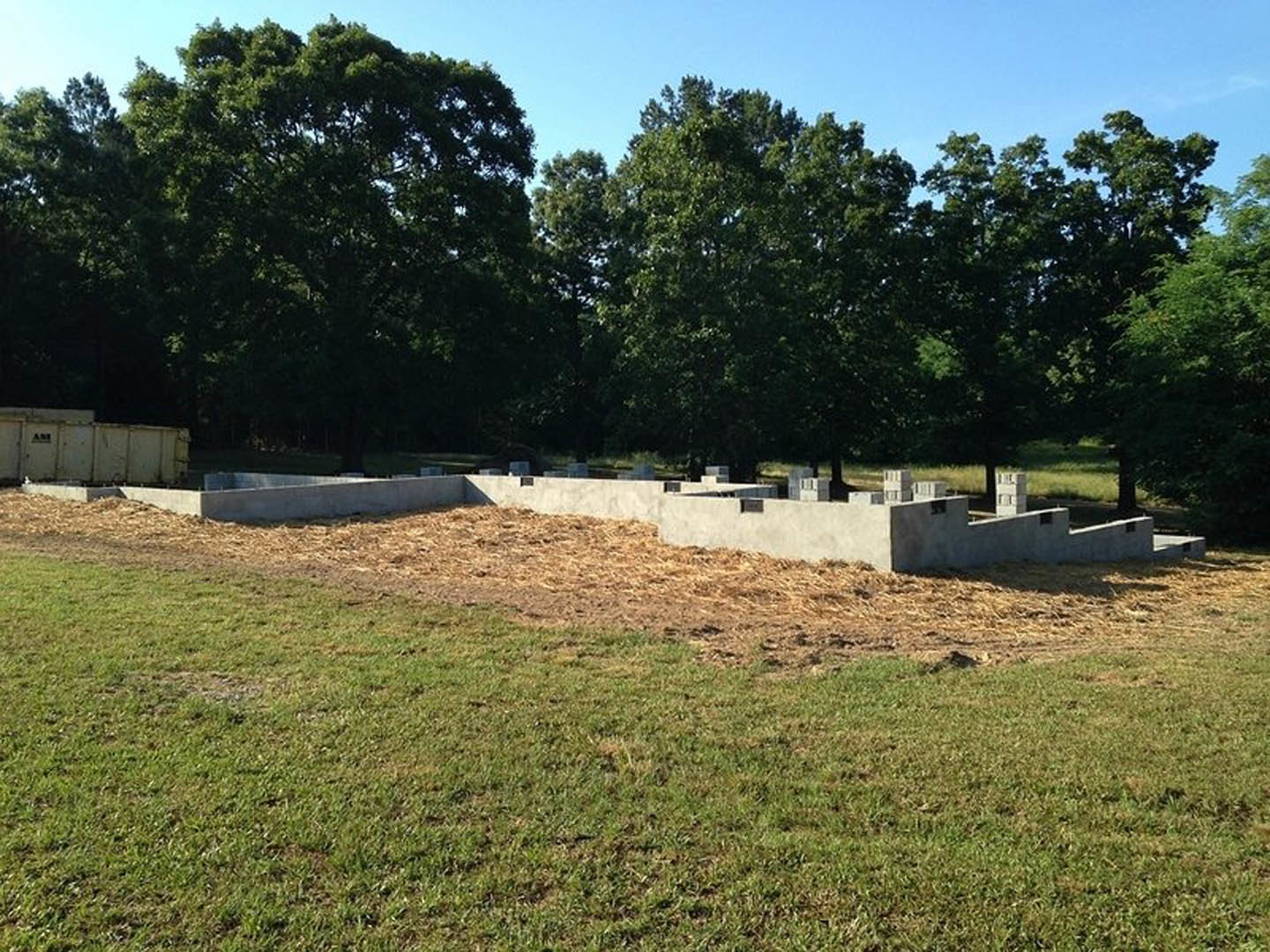 Grassy building site with scattered concrete blocks, dense trees lining the background, and a partially constructed white wall.