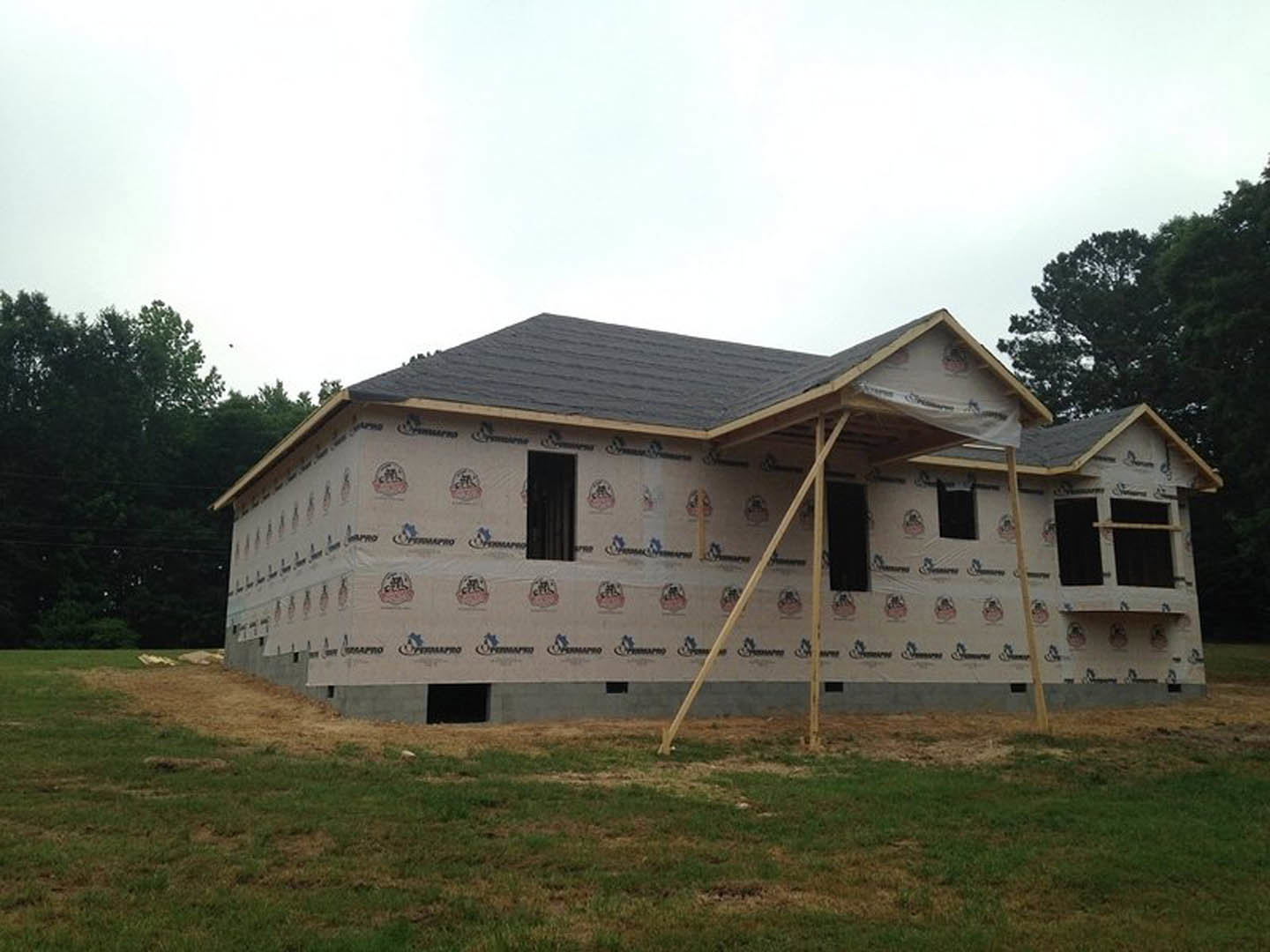 Wood-framed house under construction with covered porch, exposed roof trusses, grassy yard, and unfinished exterior walls