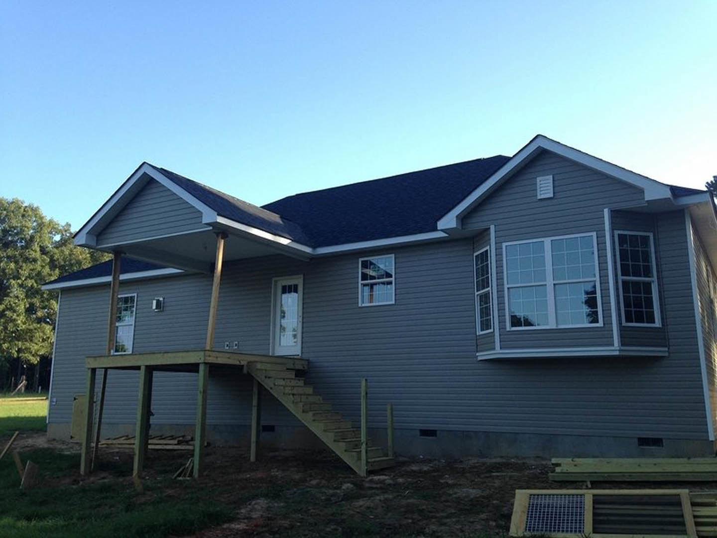 Two-story house with gray siding, white-trimmed windows, covered front porch, wooden stairs leading to entry, landscaped yard, and mature trees in background