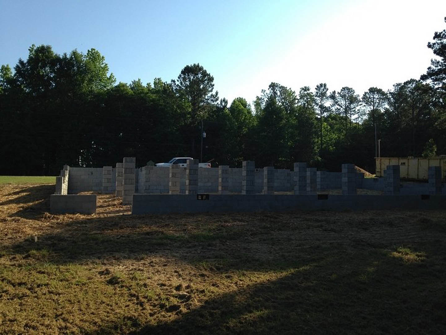 Partially built home with concrete block walls, exposed framing, and construction materials scattered on grassy lot; mature trees and parked car in background under clear blue sky.