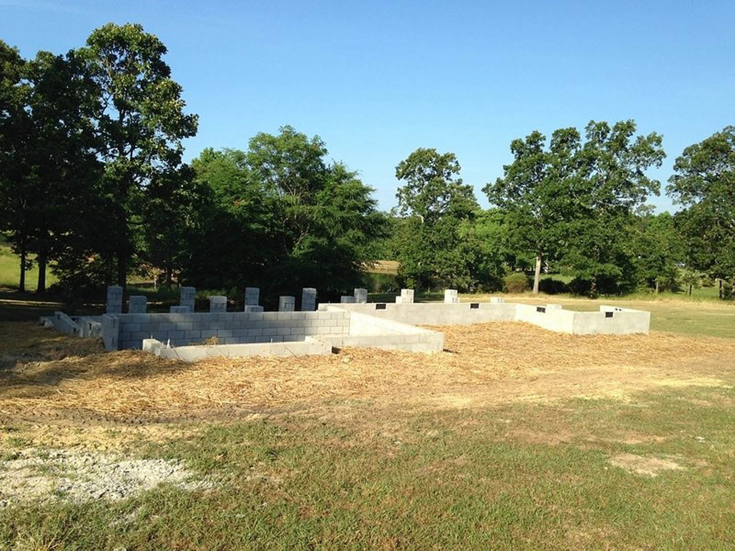 Grassy building site bordered by concrete blocks and a low concrete wall, scattered rocks in foreground, group of people standing near mature trees under blue sky.