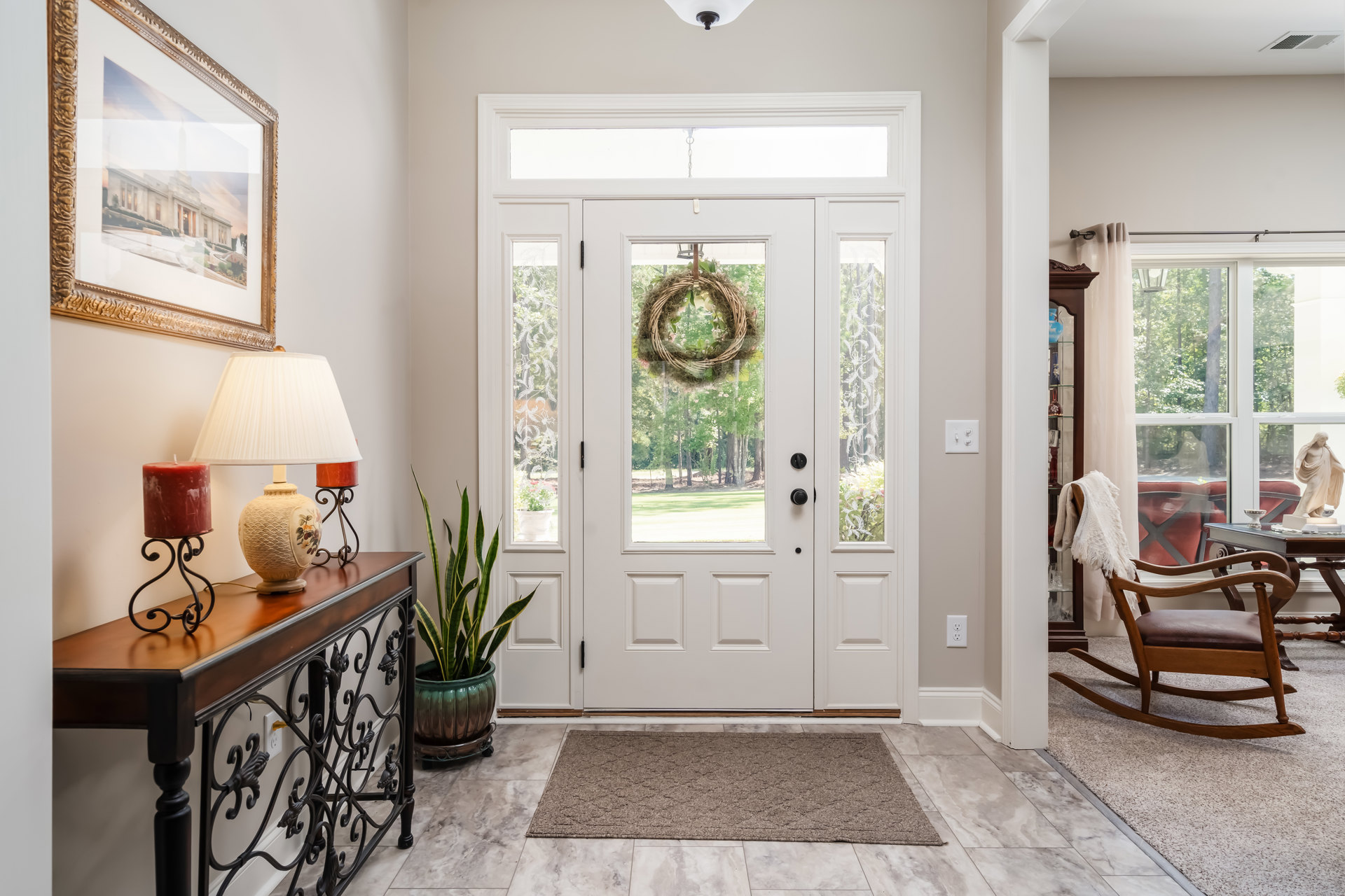 White front door with a decorative wreath, rocking chair with white fringe, side table holding a patterned vase, potted plant, framed picture on the wall