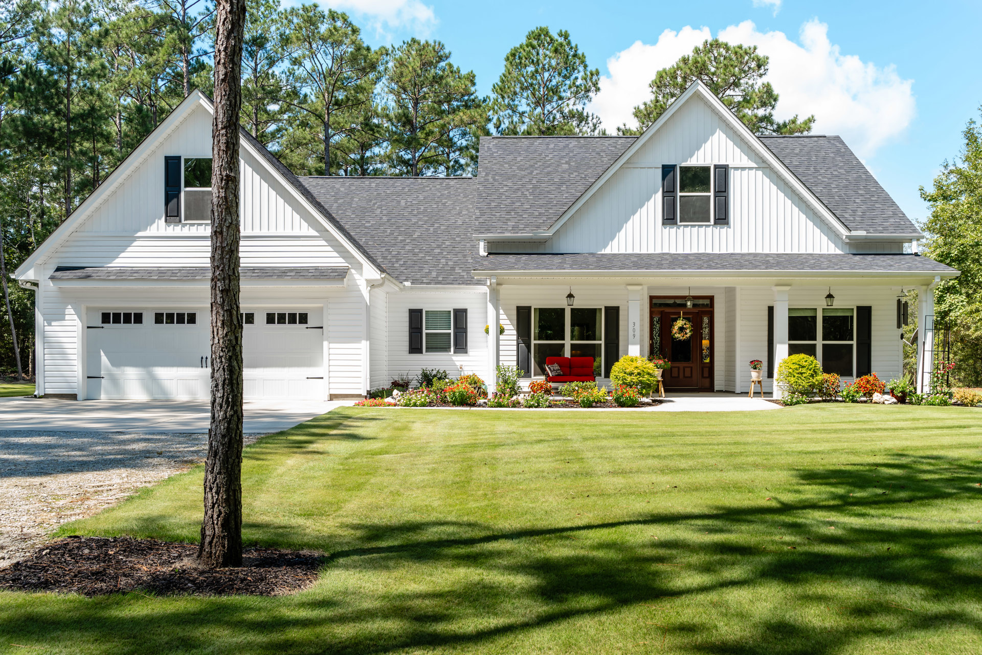 White siding house with black shuttered windows, front porch featuring a red couch with pillow, wreath-adorned door, green lawn bordered by flowers, mature trees in background