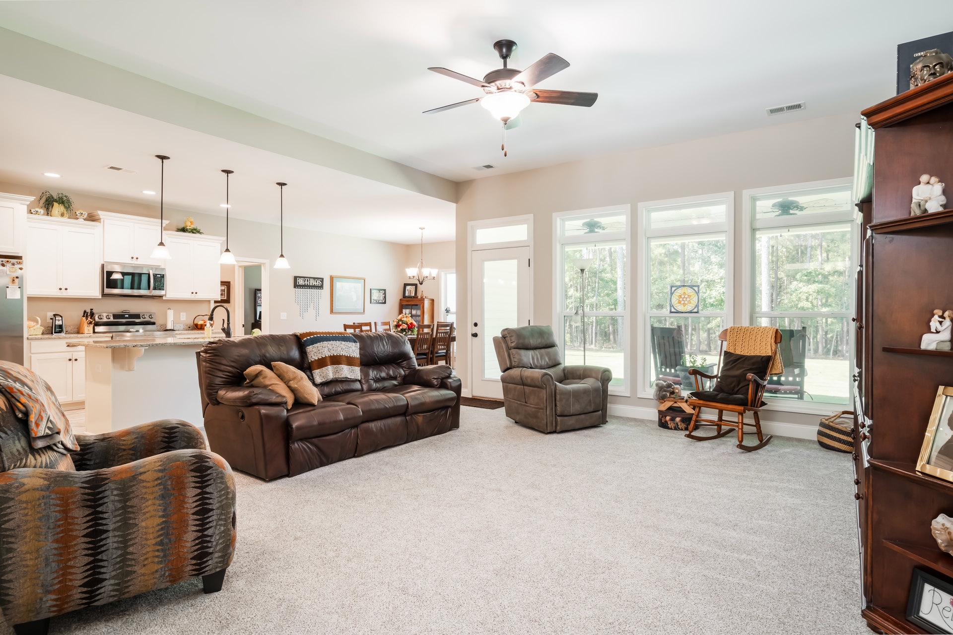Brown leather couch with pillows and a blanket, ceiling fan overhead, rocking chair with a blanket, stained glass window featuring a flower design, hardwood floors, neutral walls.