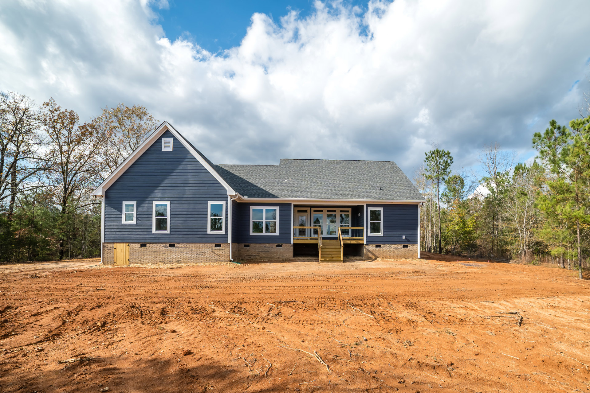 Two-story house under construction with white-framed windows, covered porch, and unfinished dirt yard featuring tire tracks; blue sky with scattered clouds reflected in window