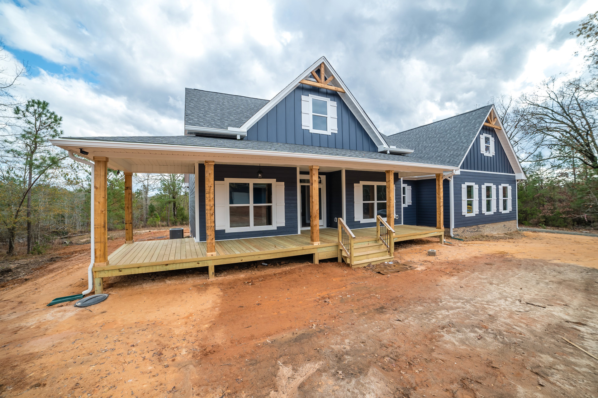 Two-story house with blue and white siding, covered front porch, white window shutters, wooden deck and stairs, triangular roof, surrounded by trees and cloudy sky.