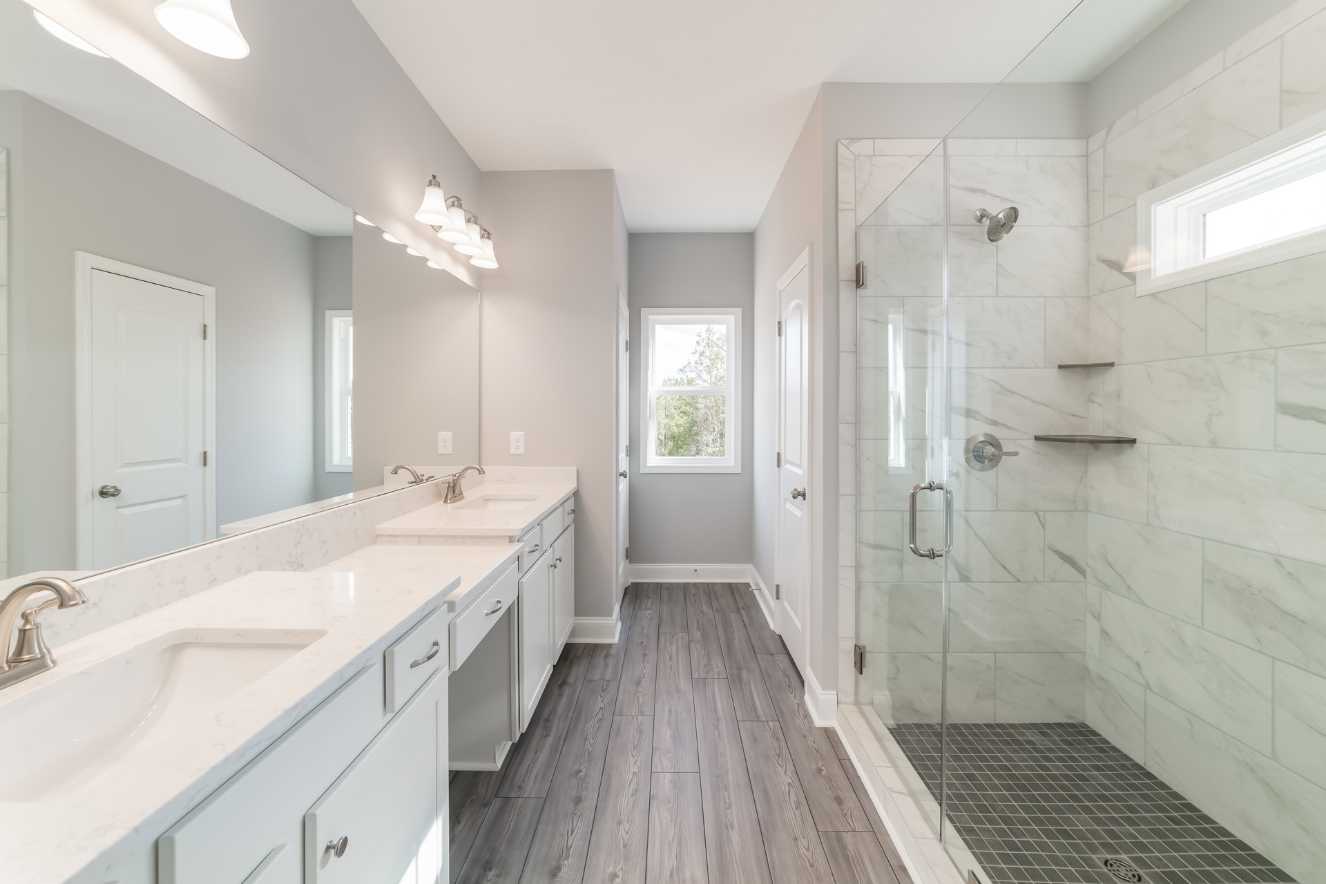 Bathroom featuring marble countertops, glass shower enclosure, large mirror, white cabinetry, silver hardware, and window overlooking trees.