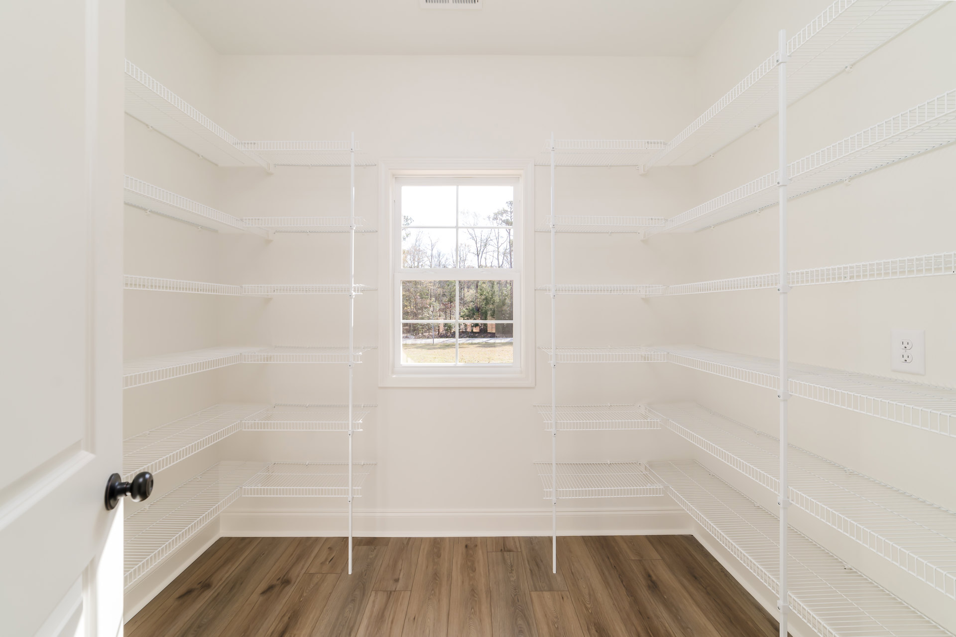 White built-in shelves along one wall, large window with view of green trees, light wood flooring with white baseboard trim, white plaster ceiling with crown molding, close-up of
