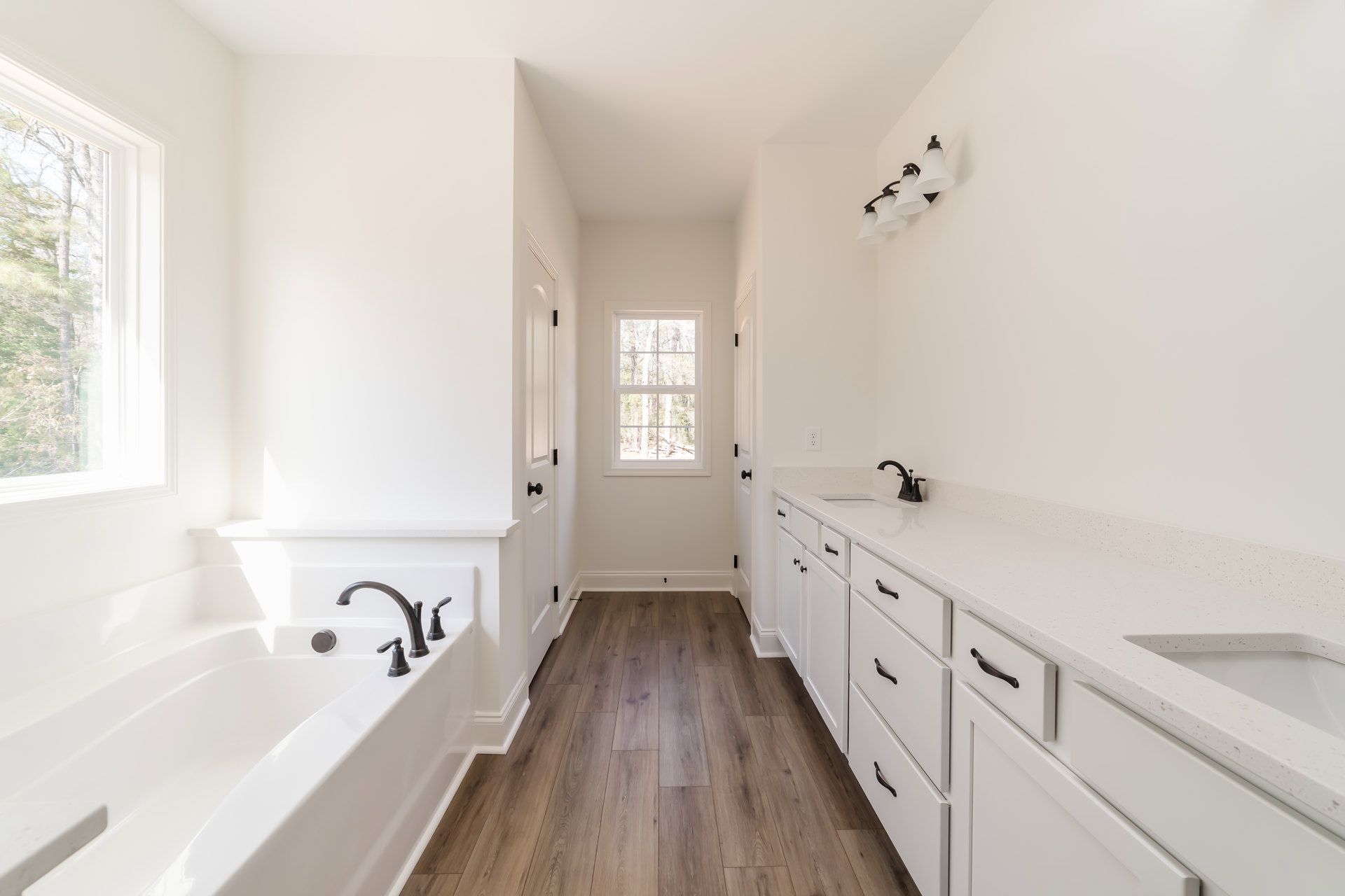Bathroom with white cabinets and white countertops, black faucet on white bathtub, four-lamp light fixture, window showing trees outside, black handles on cabinetry