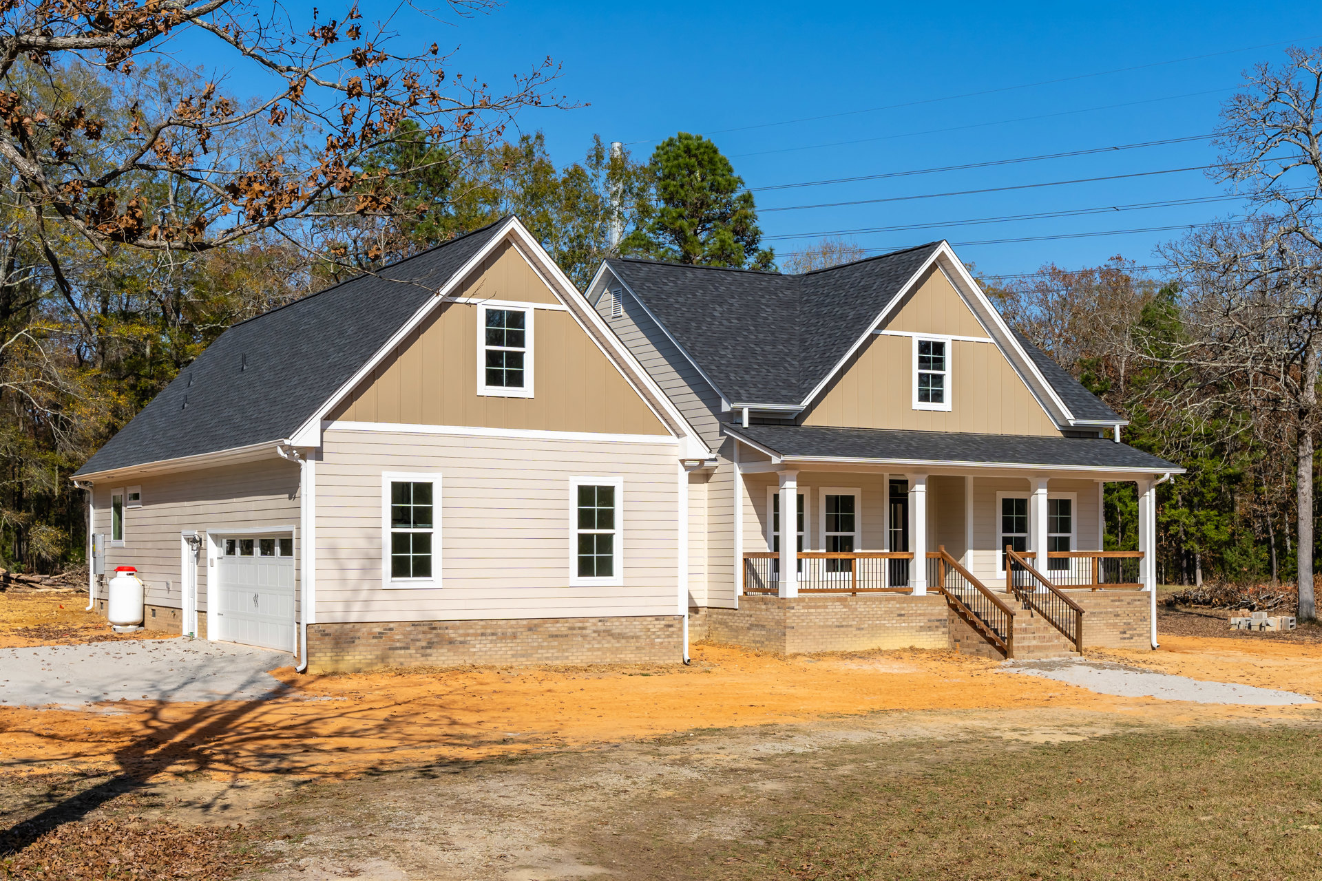Two-story house with gray siding, white-framed windows, attached garage with white door and upper windows, concrete driveway, overhead power lines, and dirt landscaping adjacent to