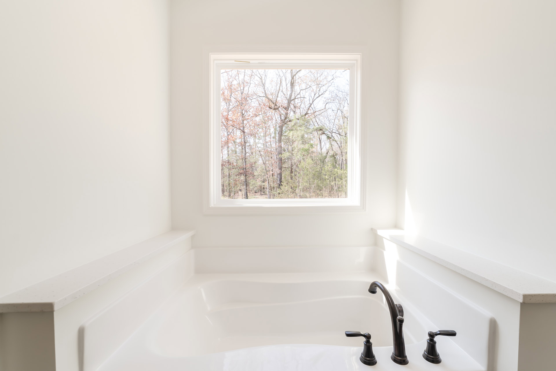 Freestanding white bathtub beneath a large window with tree views, matte plaster walls, chrome faucet, and black bell resting on tub ledge