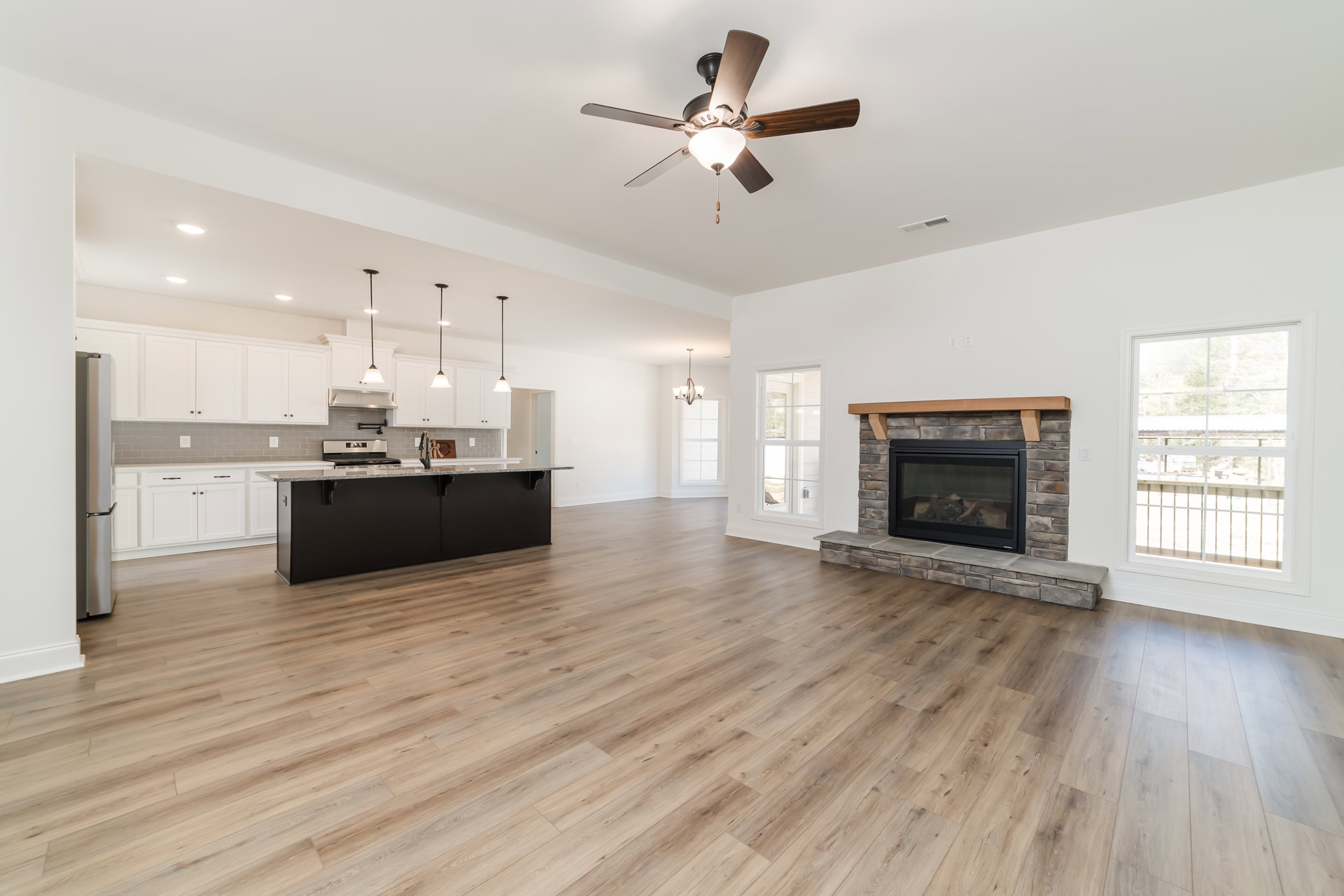 Living room with hardwood floors, ceiling fan with light fixture, glass door fireplace, kitchen counter and sink visible, window overlooking exterior railing