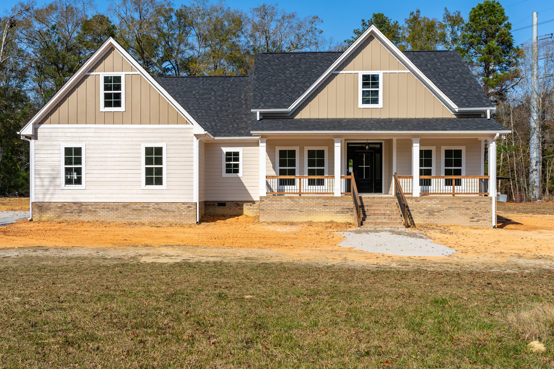 Two-story home under construction with white-framed windows, black glass-paneled door, covered porch with railing, unfinished lawn, and surrounding trees under a blue sky