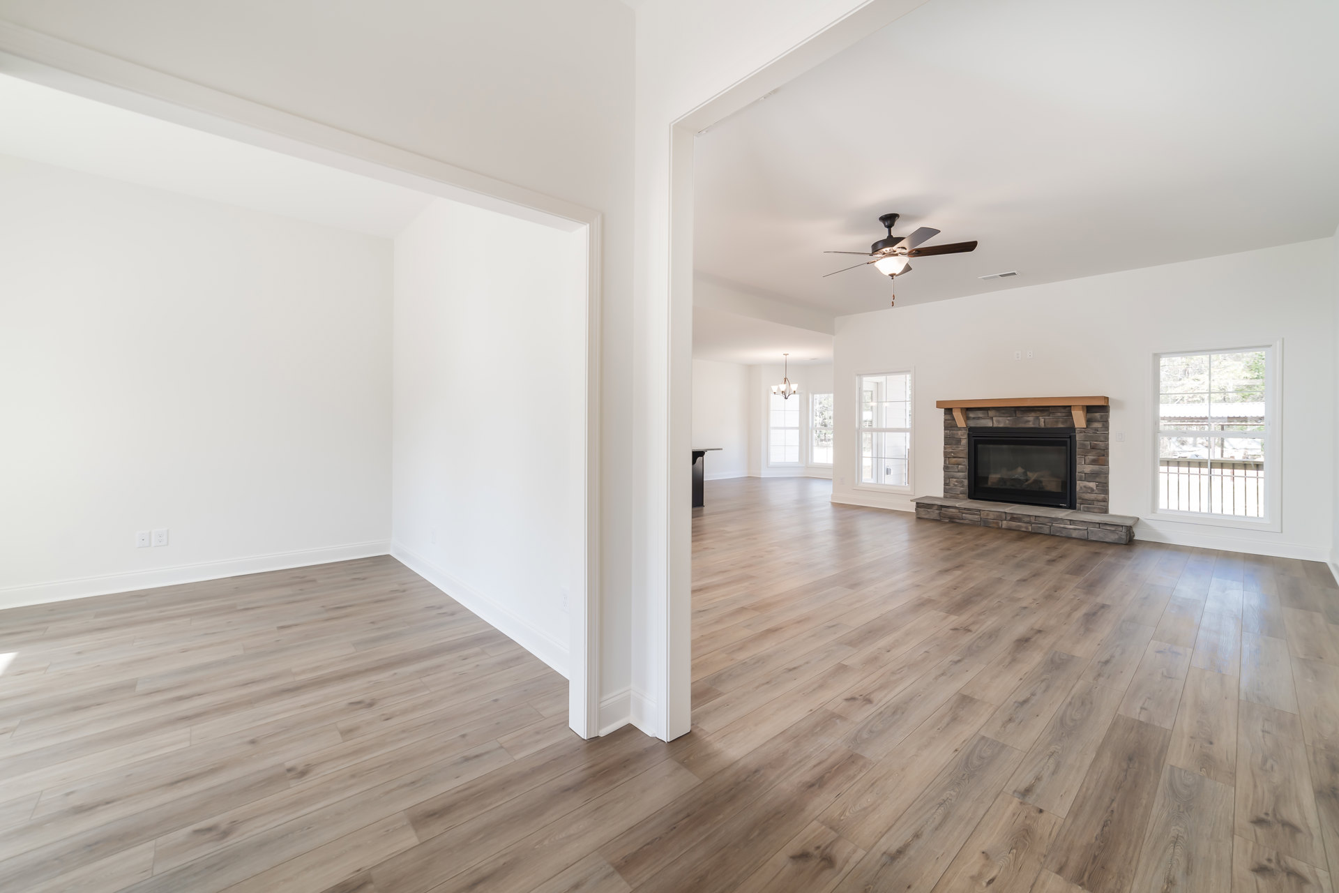 Living room featuring hardwood floors, white plaster walls, glass-door fireplace, ceiling fan with light, large window with railing overlooking trees