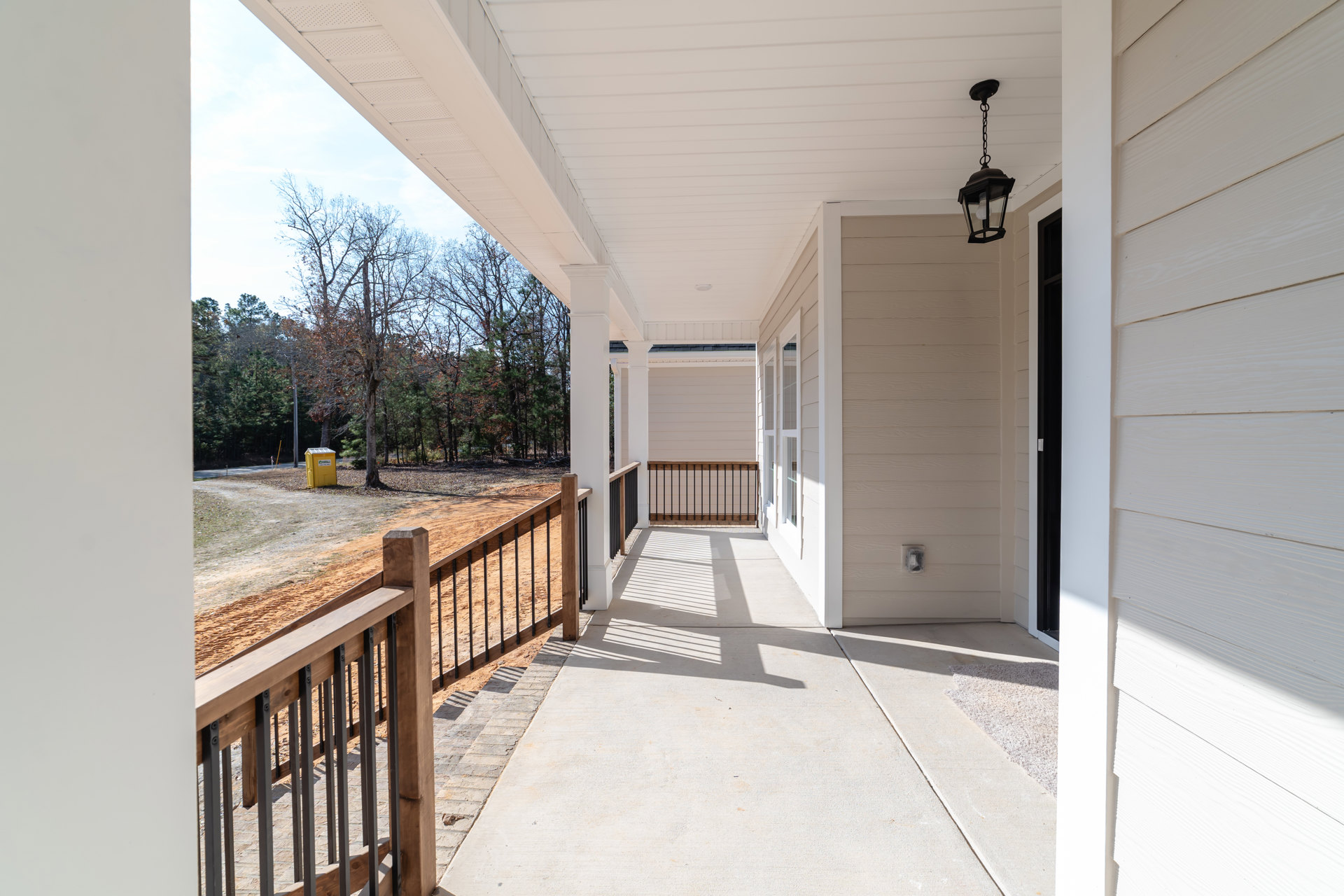 Wooden porch with black metal railing, illuminated lamp fixture, composite deck flooring, tree and fence visible in background.