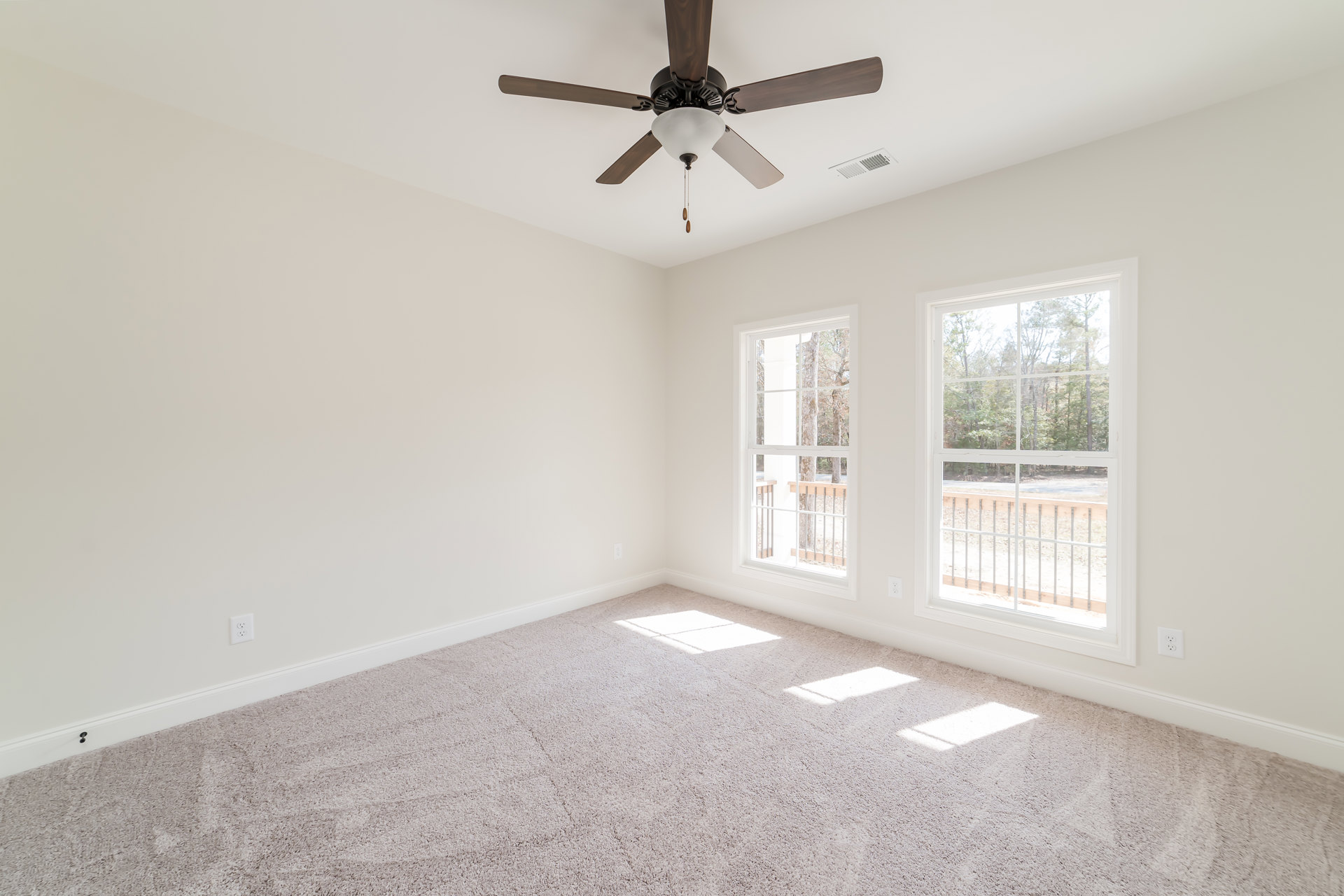 Ceiling fan with light fixture mounted on white plaster ceiling, large windows with view of trees, carpeted floor, sunlight casting square shadow on ground.