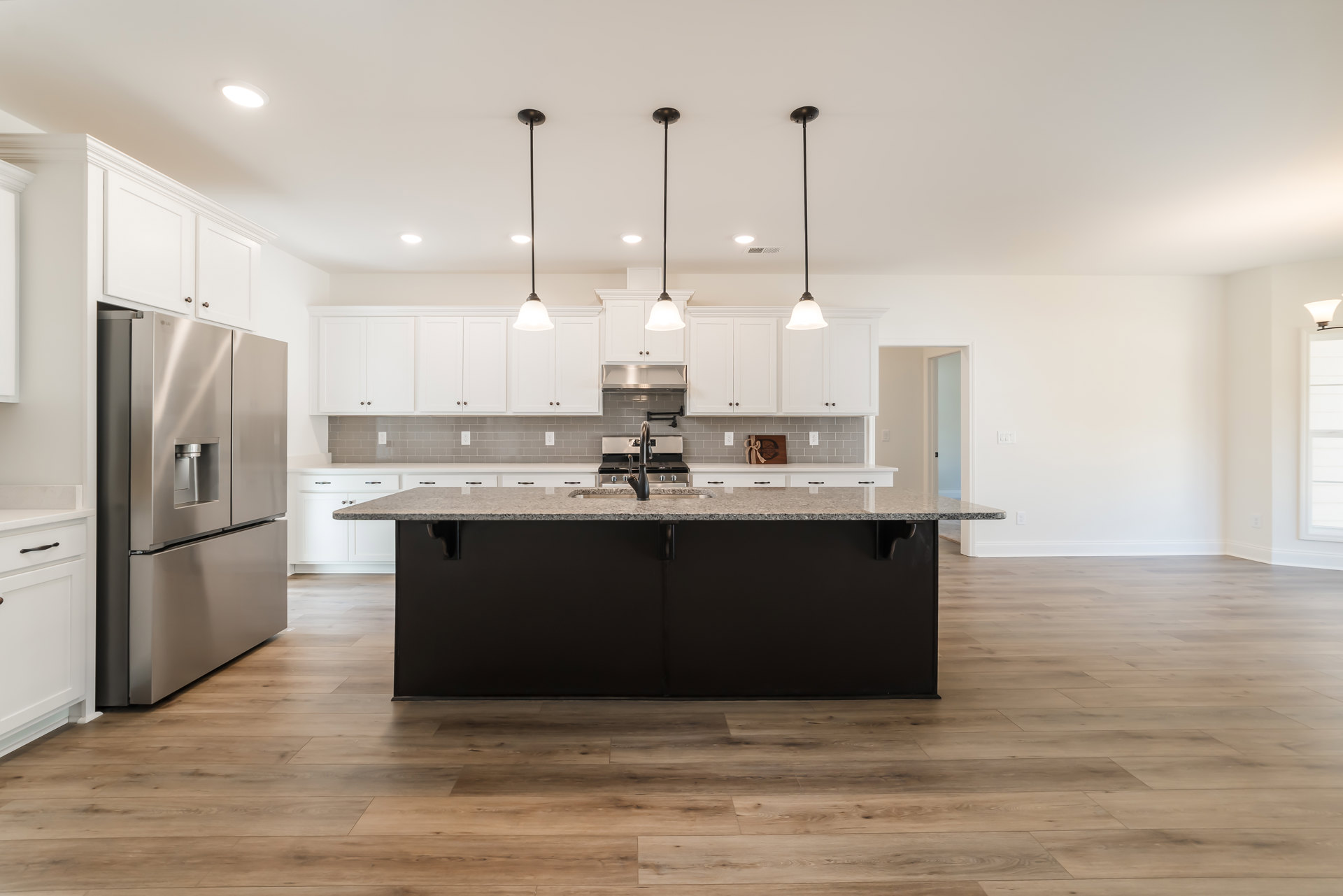 Spacious kitchen featuring a large central island with stone countertop, tile flooring, white cabinetry, stainless steel refrigerator, built-in sink, and modern pendant lighting