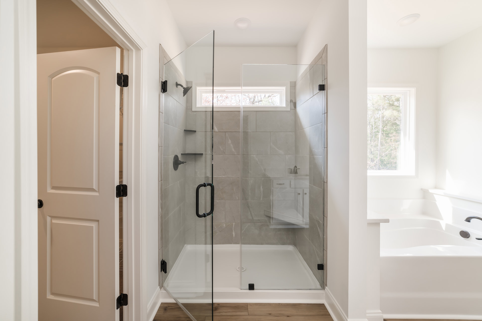 Glass shower enclosure with chrome hardware, white tile walls, window overlooking trees, white door with black hinges and handle, modern bathroom fixtures