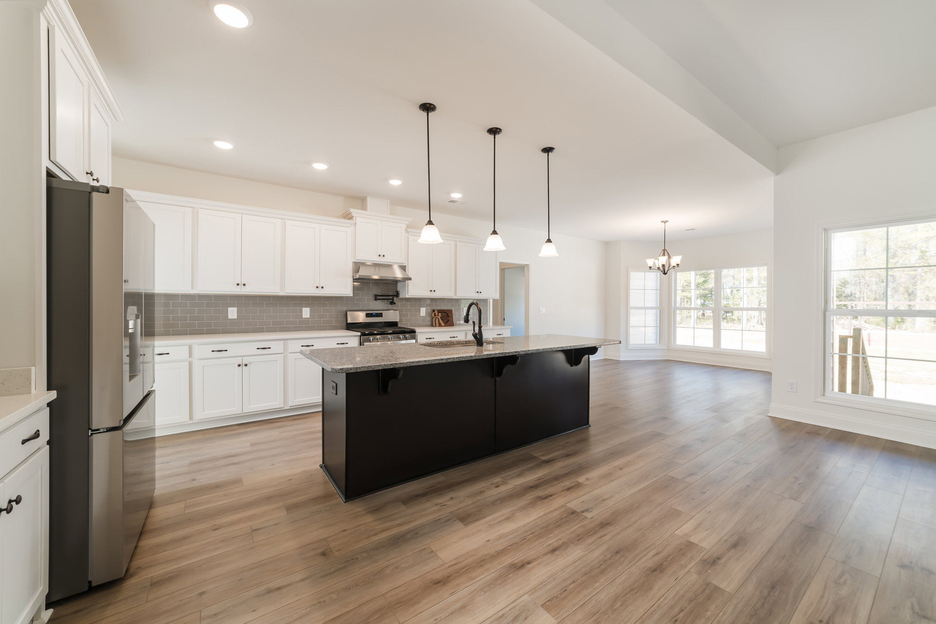 Spacious kitchen featuring a large central island with built-in sink, white cabinetry, stainless steel refrigerator, wood flooring, and a window overlooking a wood staircase with