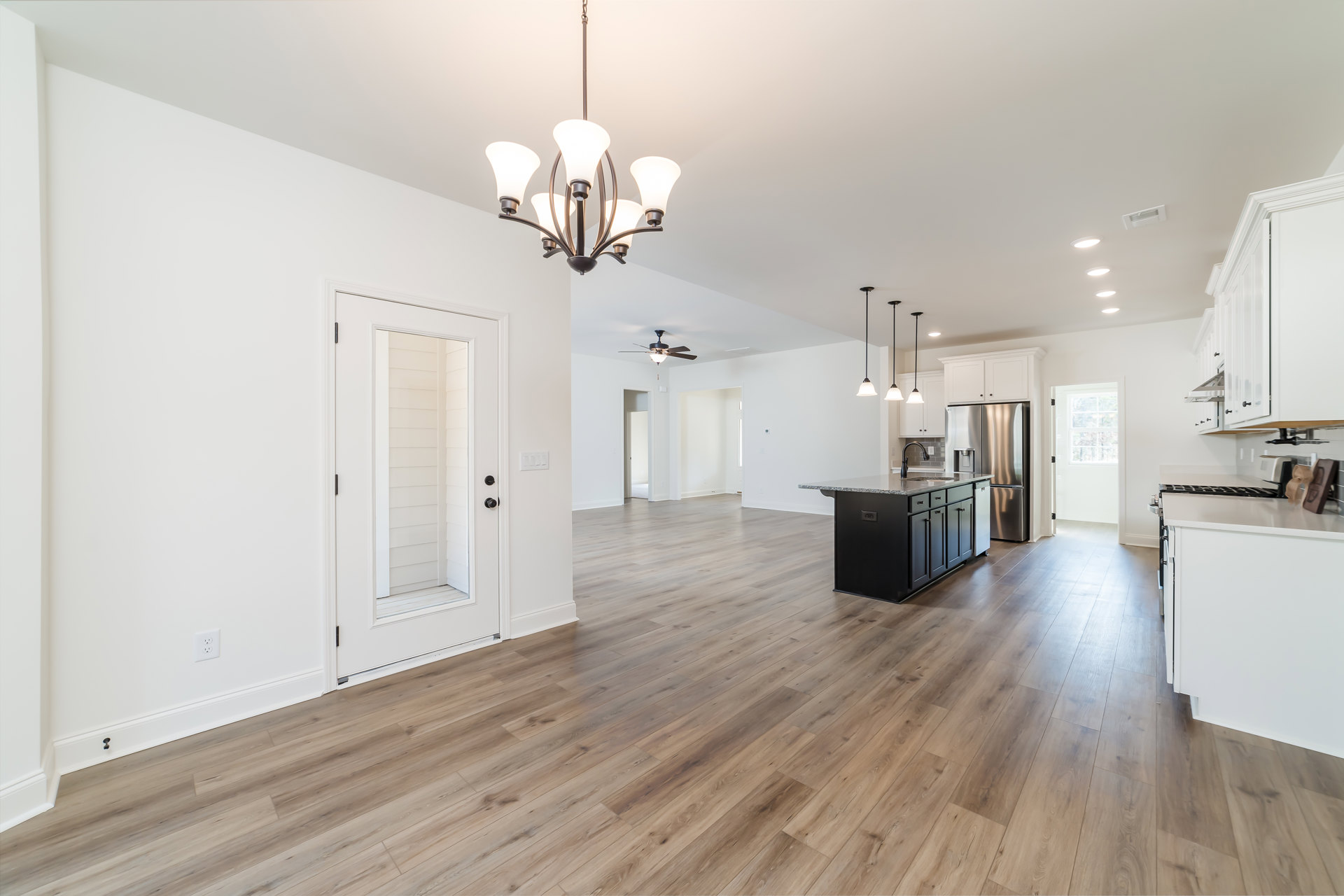Spacious open concept kitchen and living area featuring a central kitchen island with sink, stainless steel refrigerator, hardwood flooring, white walls, and a modern chandelier.
