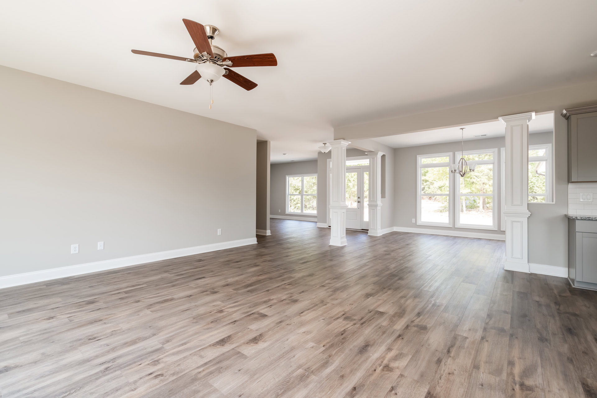 Ceiling fan with light fixture, hardwood floor, white pillars, large window with view of trees, neutral walls