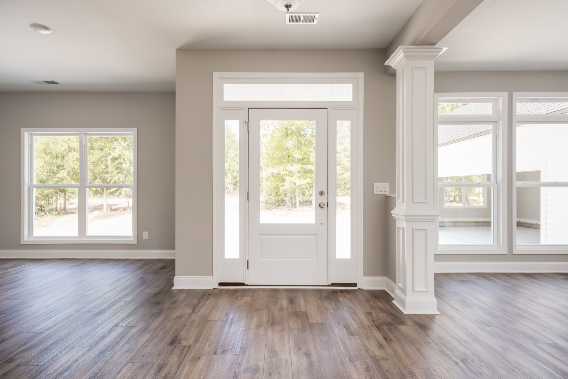 White door with glass panes, wood flooring, window showing trees outside, ceiling vent, and light fixture in a bright room.