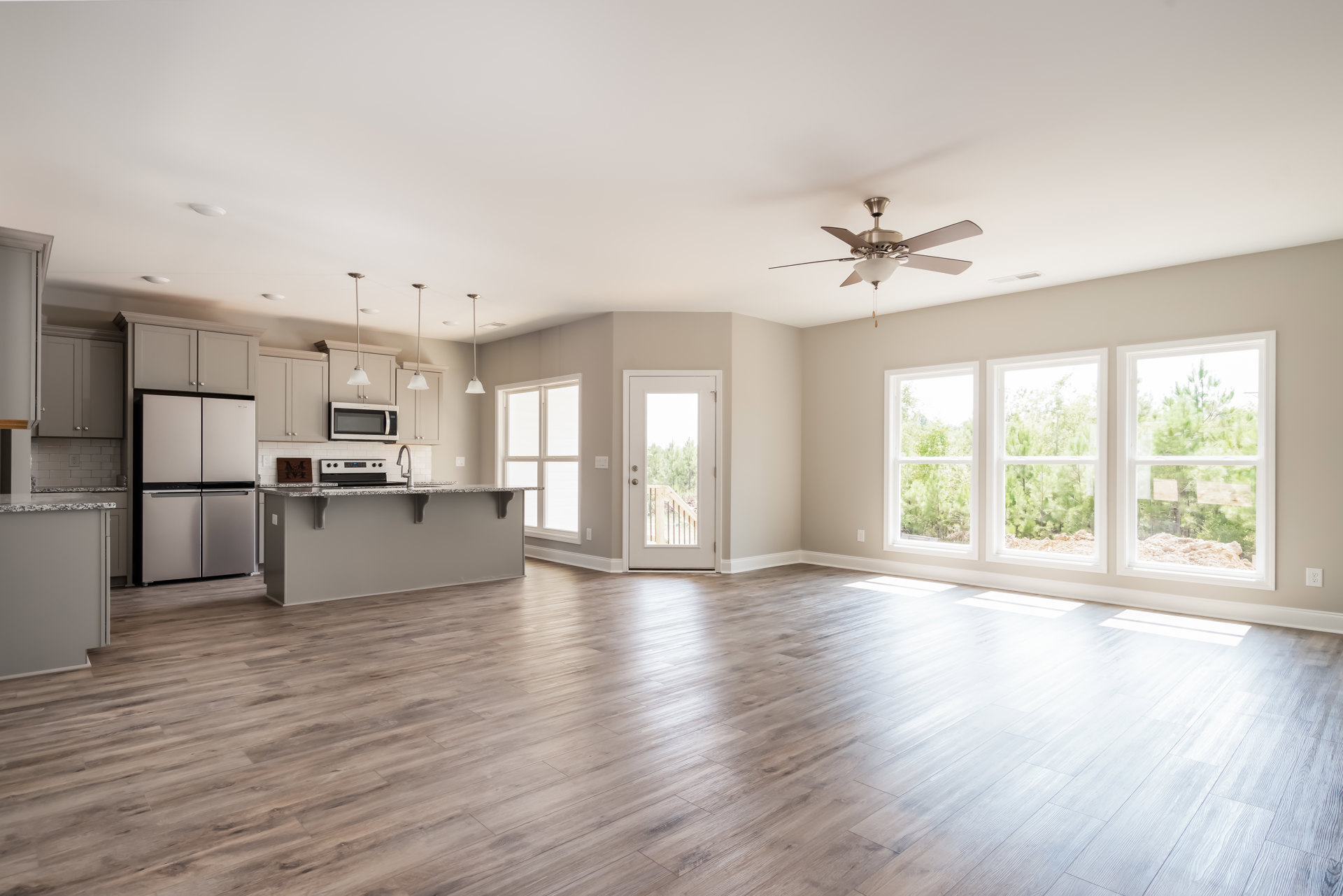 Open-concept kitchen and living room with hardwood flooring, white cabinetry, stainless steel microwave, white refrigerator with black handles, row of windows showing trees