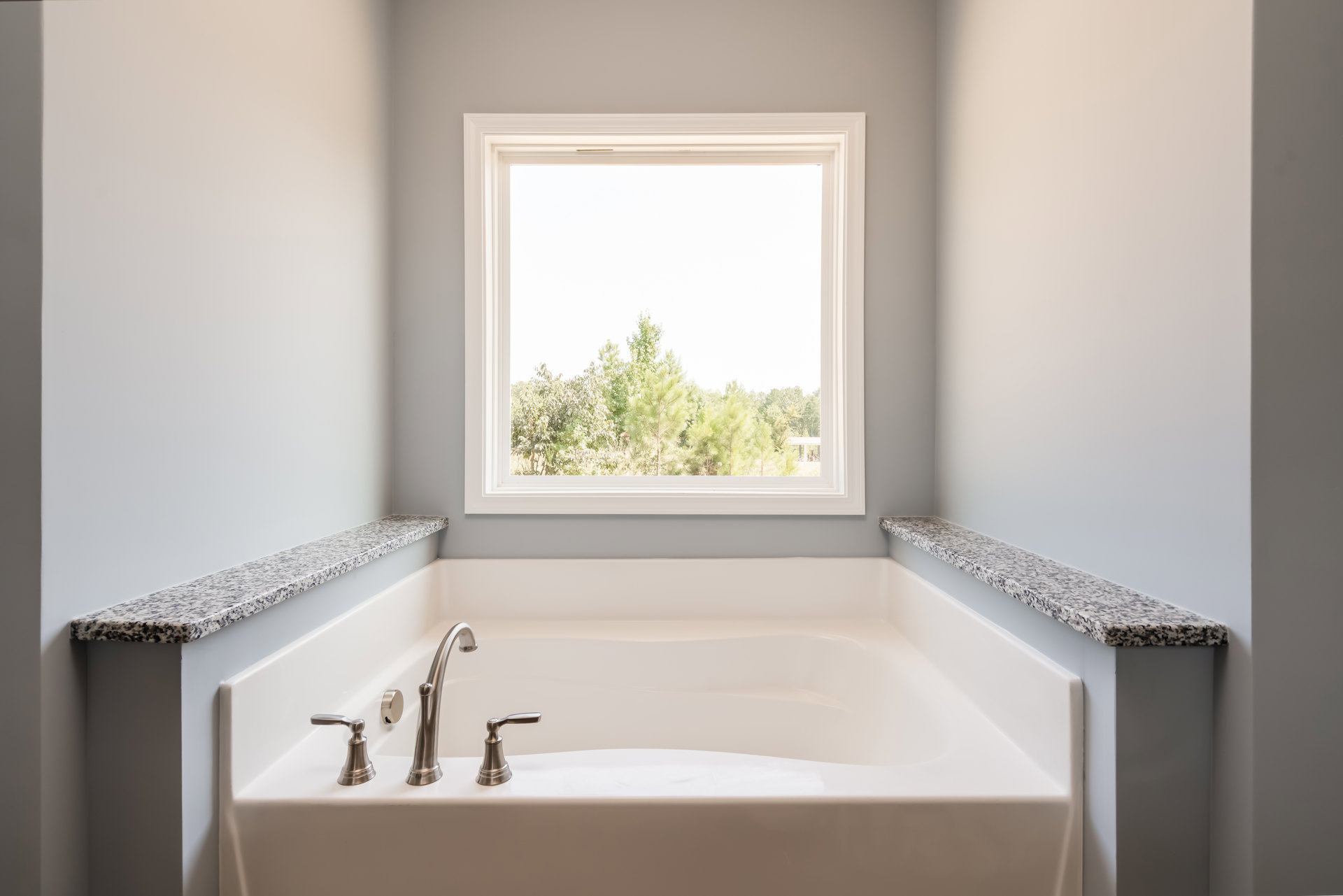Freestanding white bathtub beneath large window with tree views, chrome faucet, light-colored walls, and stone countertop visible