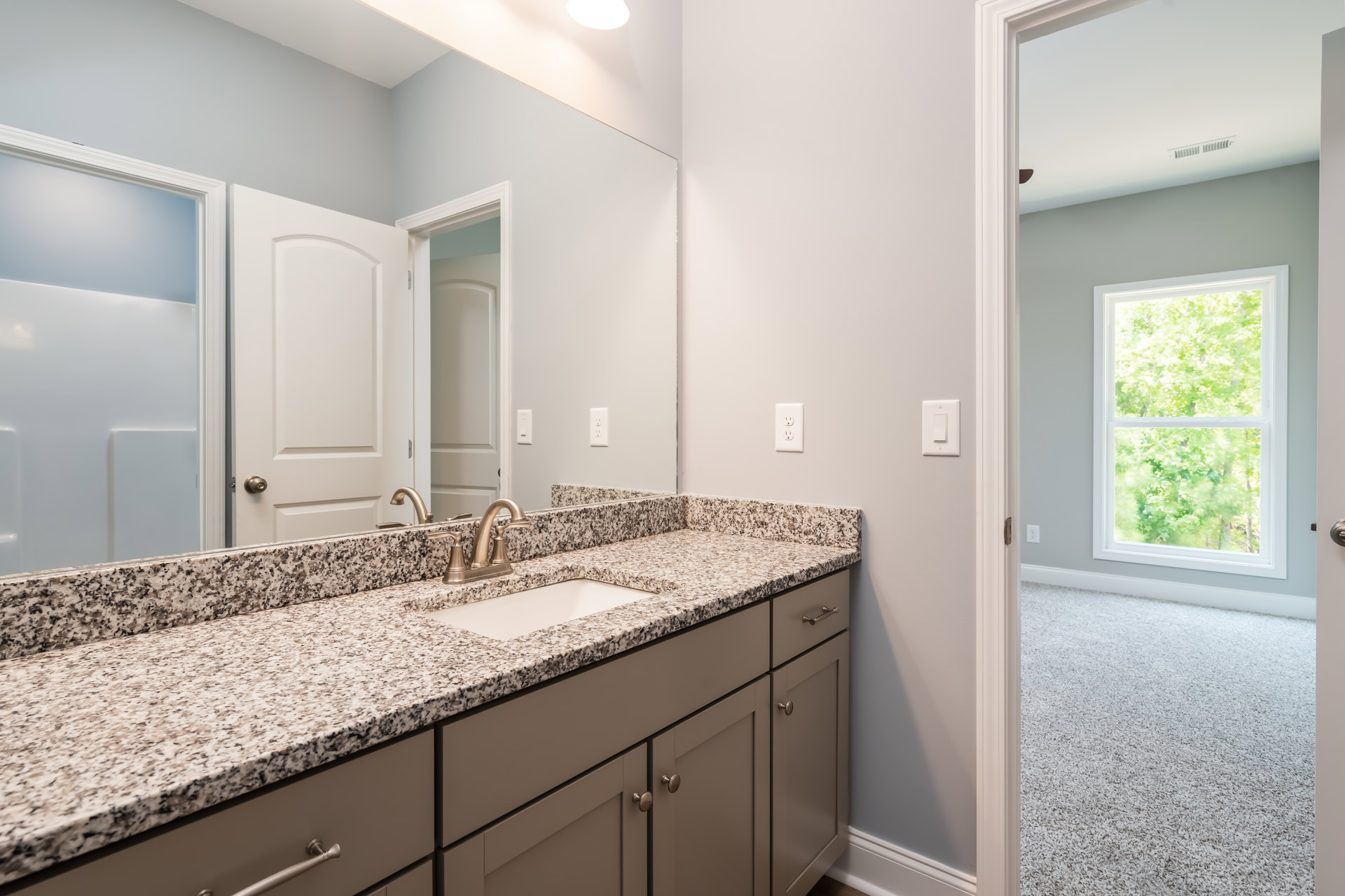 Granite bathroom countertop with undermount sink, white cabinetry, chrome faucet, tile backsplash, window overlooking trees, white door with metal handle, and wall-mounted light
