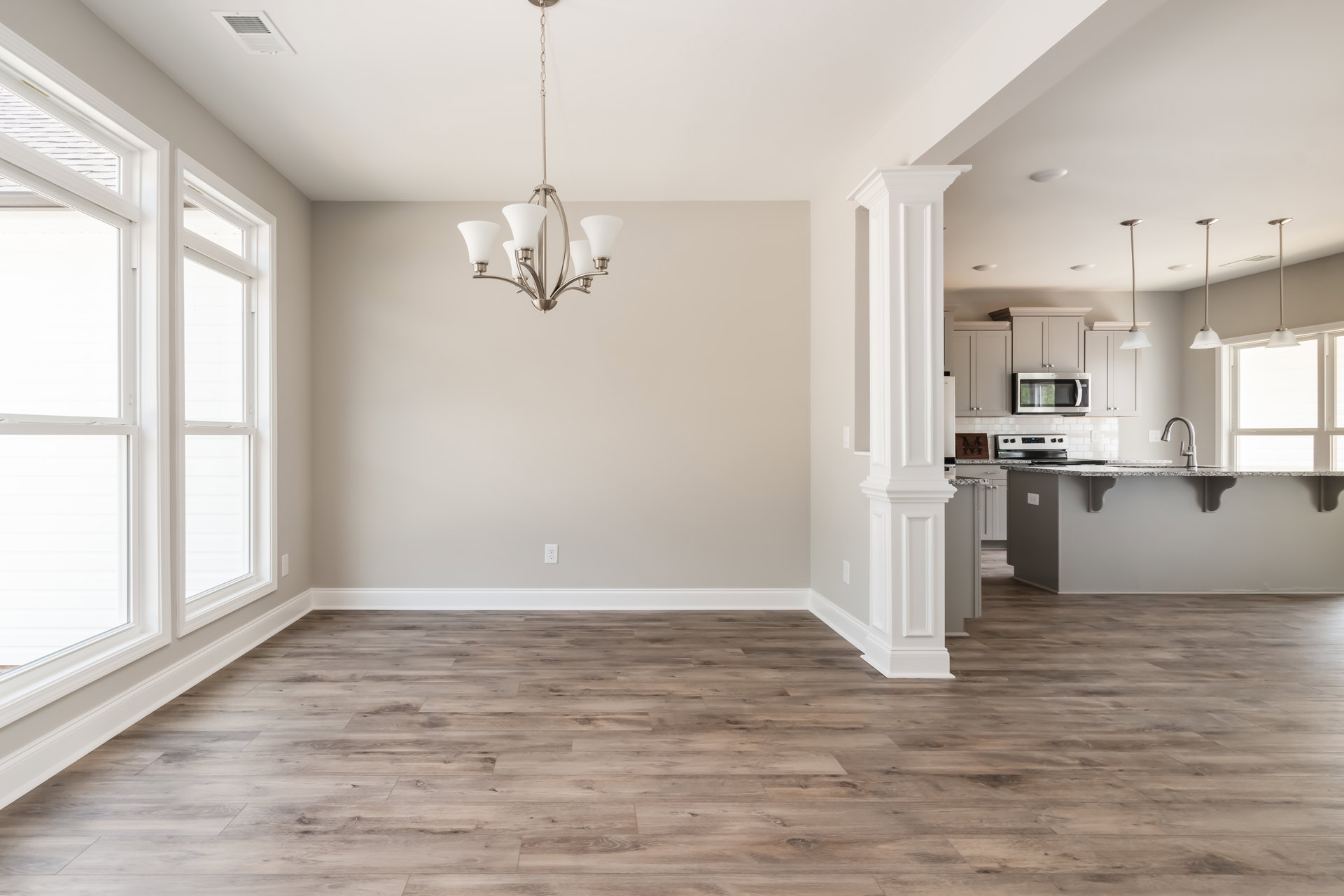 Wood flooring, white plaster walls, ornate chandelier hanging from ceiling, cabinetry along one wall, large window with trim, person standing near built-in microwave.